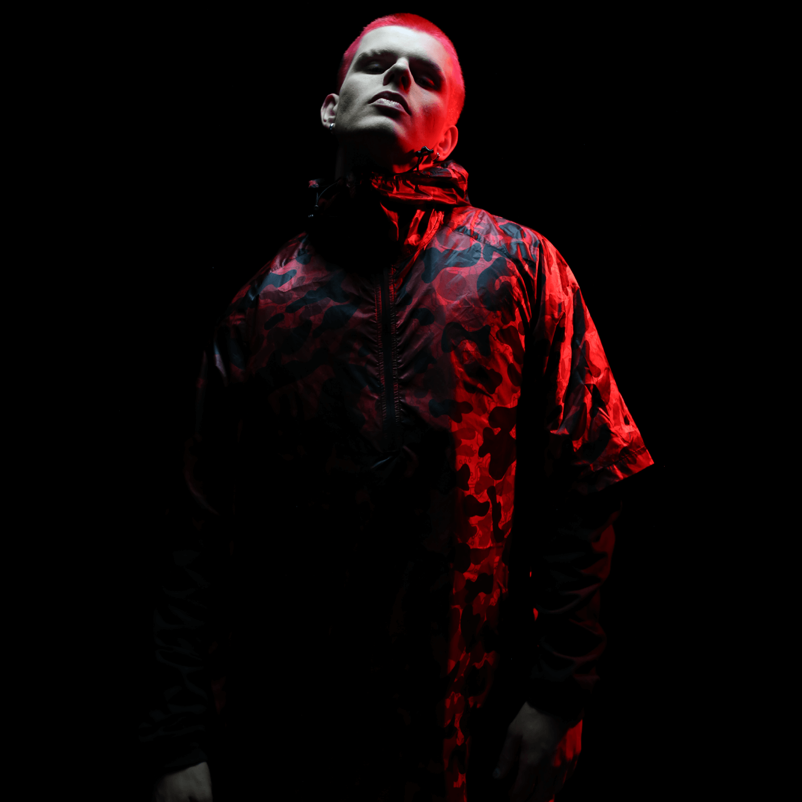 A person stands against a dark backdrop, donning a hooded jacket that features a striking red camouflage design. Dramatic lighting casts deep shadows around them, highlighting their face and the Diablo Parachute Poncho by Machina - Wearable Technology. This water-resistant poncho adds an enigmatic touch to their formidable silhouette.
