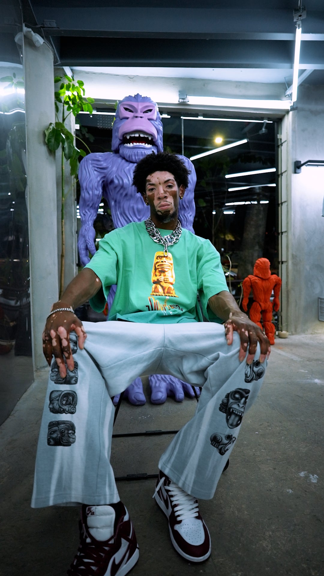 A man sits on a large purple gorilla sculpture, dressed in a mint green T-shirt featuring a glowing design by Machina - Wearable Technology and light-colored pants adorned with printed faces. The scene is bathed in overhead lighting, casting shadows reminiscent of the Quimbaya golden stone, while a small orange figure stands in the background.