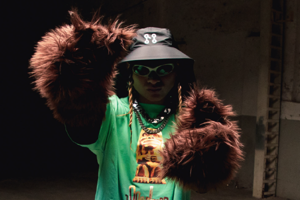 A person wearing a green shirt with furry brown sleeves strikes a confident pose, accentuated by the Explorer Hat from Machina - Wearable Technology, which doubles as a fashionable accessory. With sunglasses and layered necklaces, they stand against a dark, industrial backdrop, effortlessly blending style and weather protection.