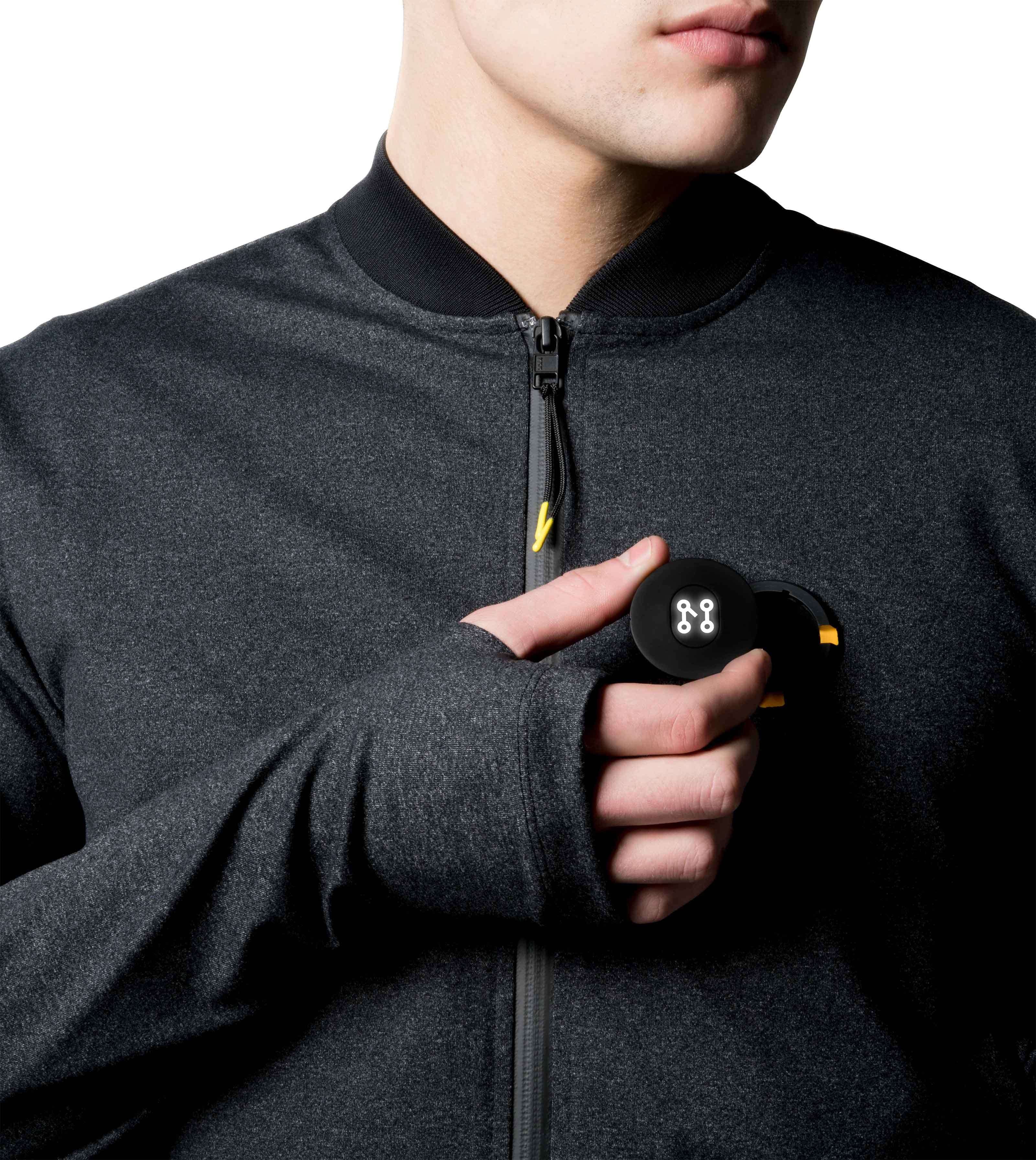 Dressed in a sleek BLACK BOMBER JACKET from Machina - Wearable Technology, a person with the jacket zipped up adjusts a small circular black device, featuring the brand's logo, attached to their chest. The person's face is partially out of frame.