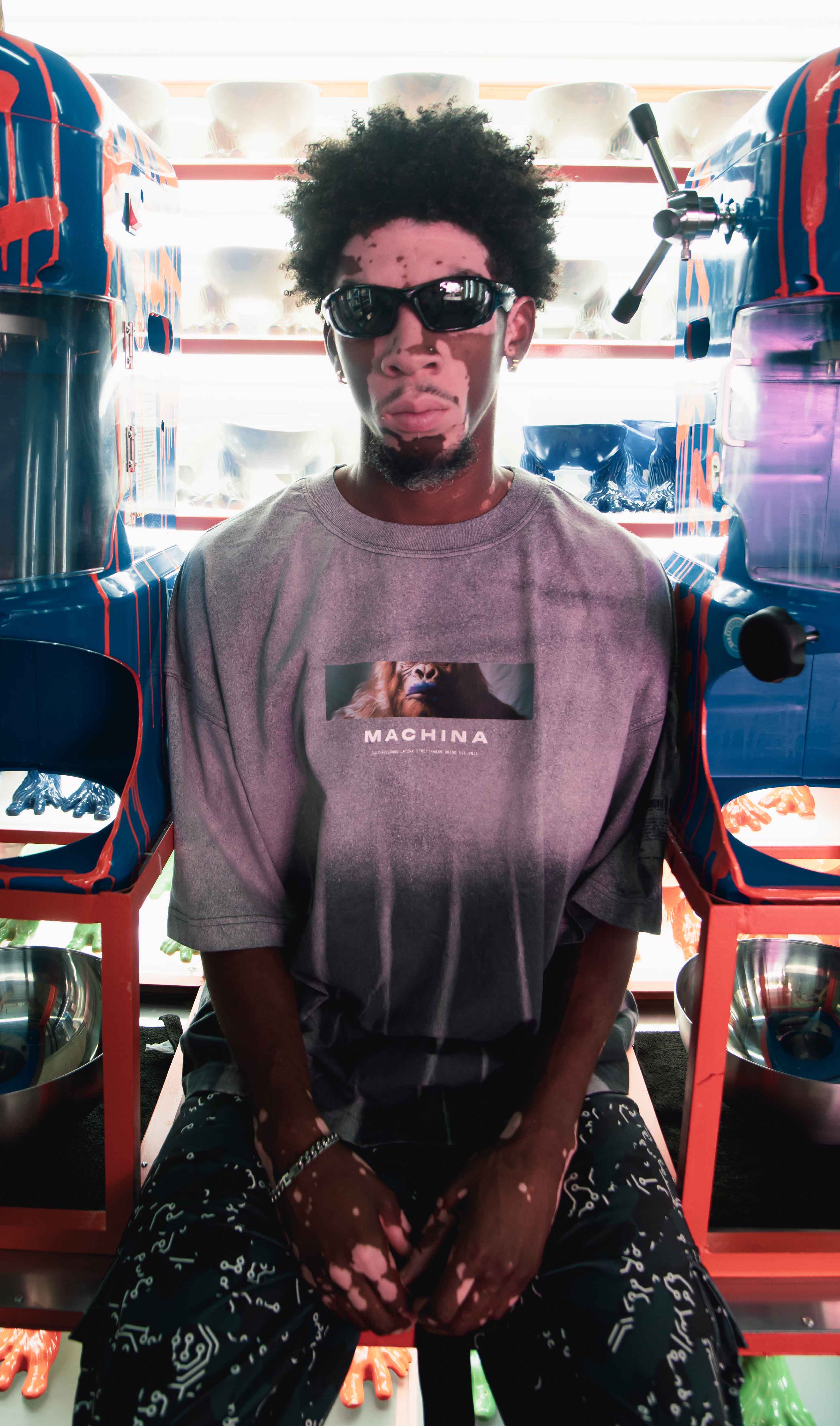 A man with curly hair and sunglasses is seated between two colorful machines. He wears a graphic T-shirt with MACHINA on it and patterned pants. Bright light shines in the background, creating a vibrant atmosphere.