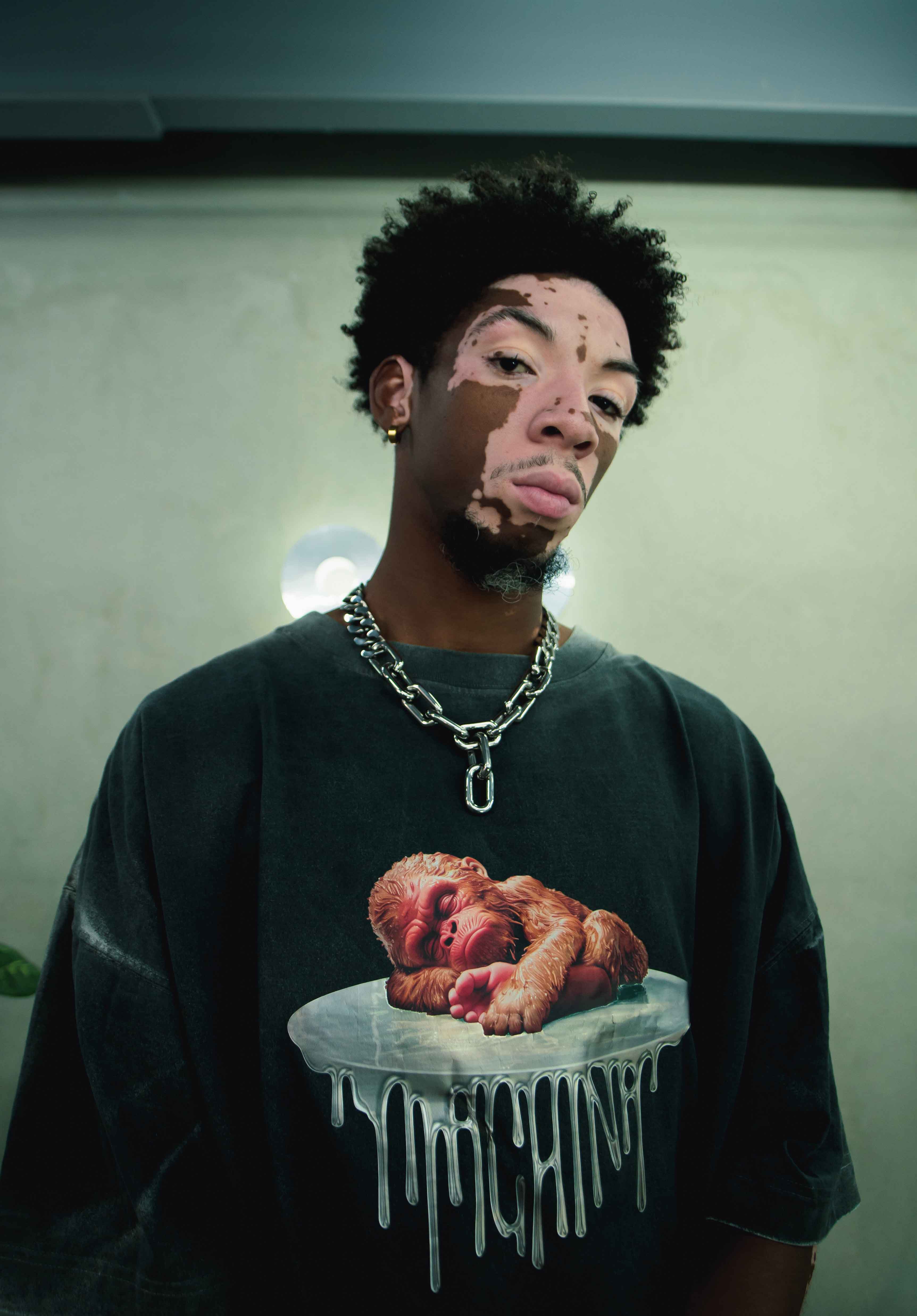 A person with vitiligo and curly hair is wearing a black "Pie Pequeño" shirt from the brand Machina - Wearable Technology, which features a graphic of a sleeping baby on a melting pedestal, suggesting an engineered cryptid. They accessorize with a heavy chain necklace while standing against a neutral background.
