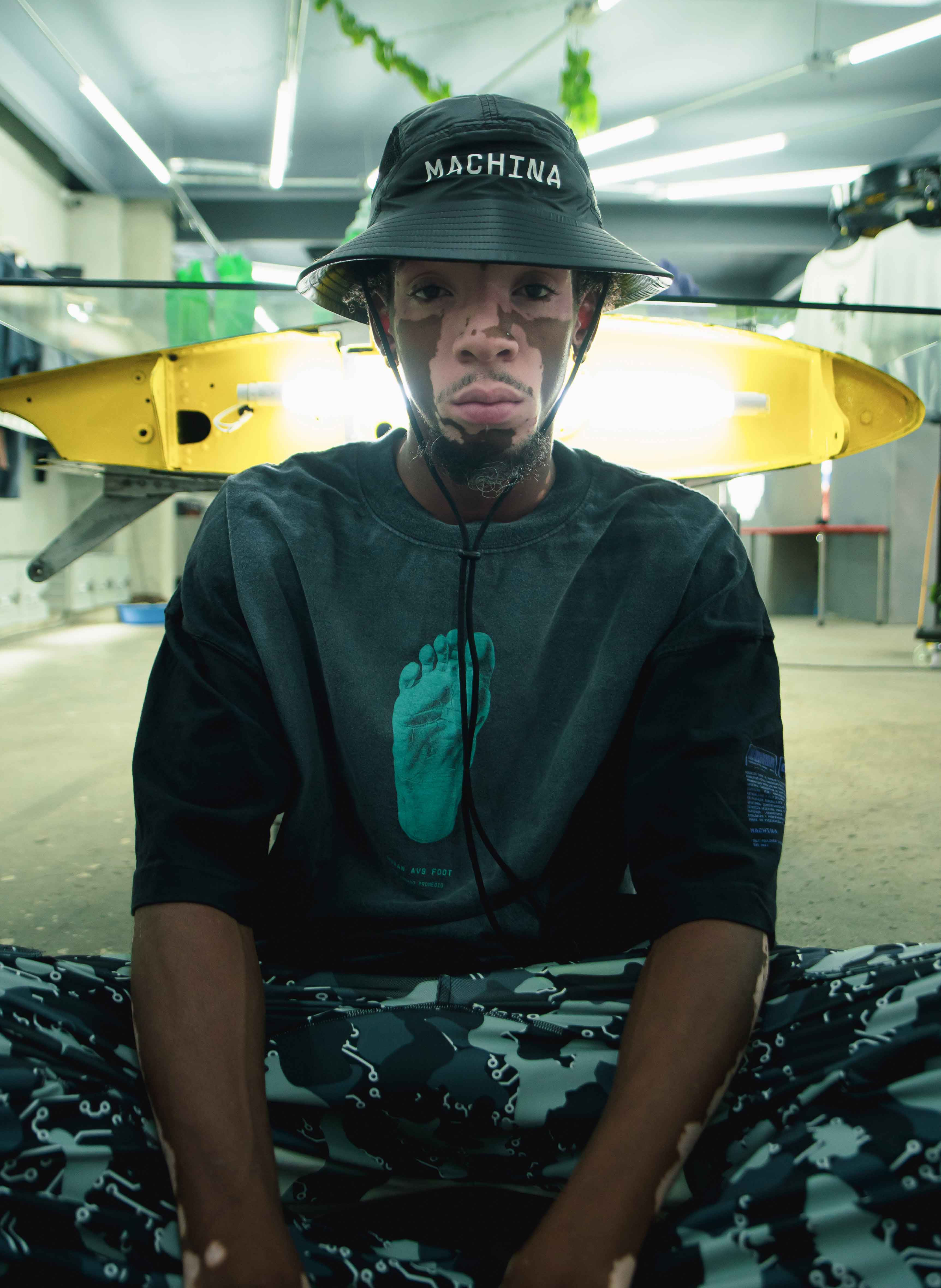 A person wearing a dark bucket hat from MACHINA - Wearable Technology and a black shirt featuring the "Footprint" design, which echoes Bigfoot's distinctive anatomy, sits cross-legged in a brightly illuminated garage. A yellow kayak or surfboard can be seen in the background, adding an intriguing blend of fashion and environment.