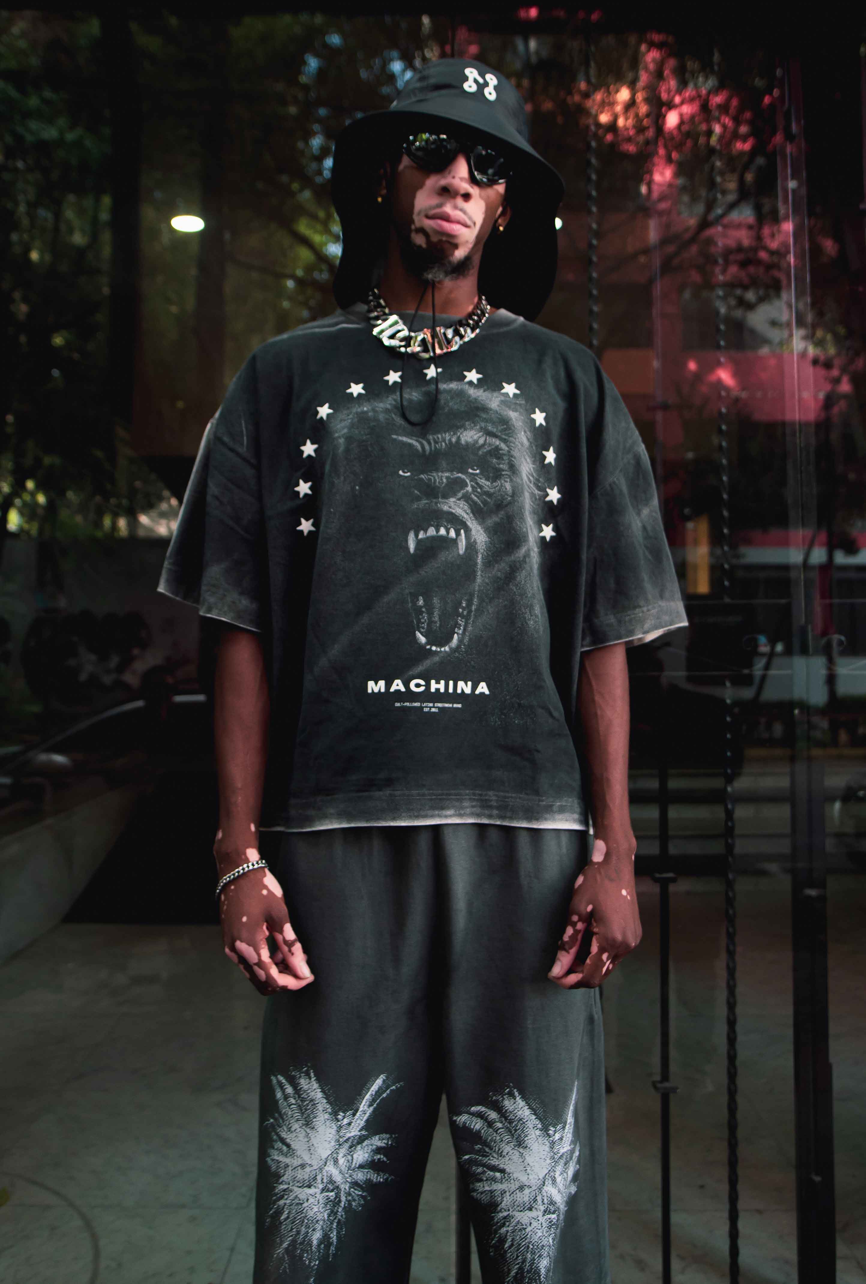 Dressed head-to-toe in the sleek ensemble from Machina - Wearable Technology, a person embodies El Protector in a stylish black outfit. They sport a wide-brimmed hat and sunglasses, while their shirt showcases a roaring gorilla surrounded by stars, symbolizing the Forest's Shadow. The look is completed with palm tree design pants, adding flair as they stand confidently in a modern urban setting.