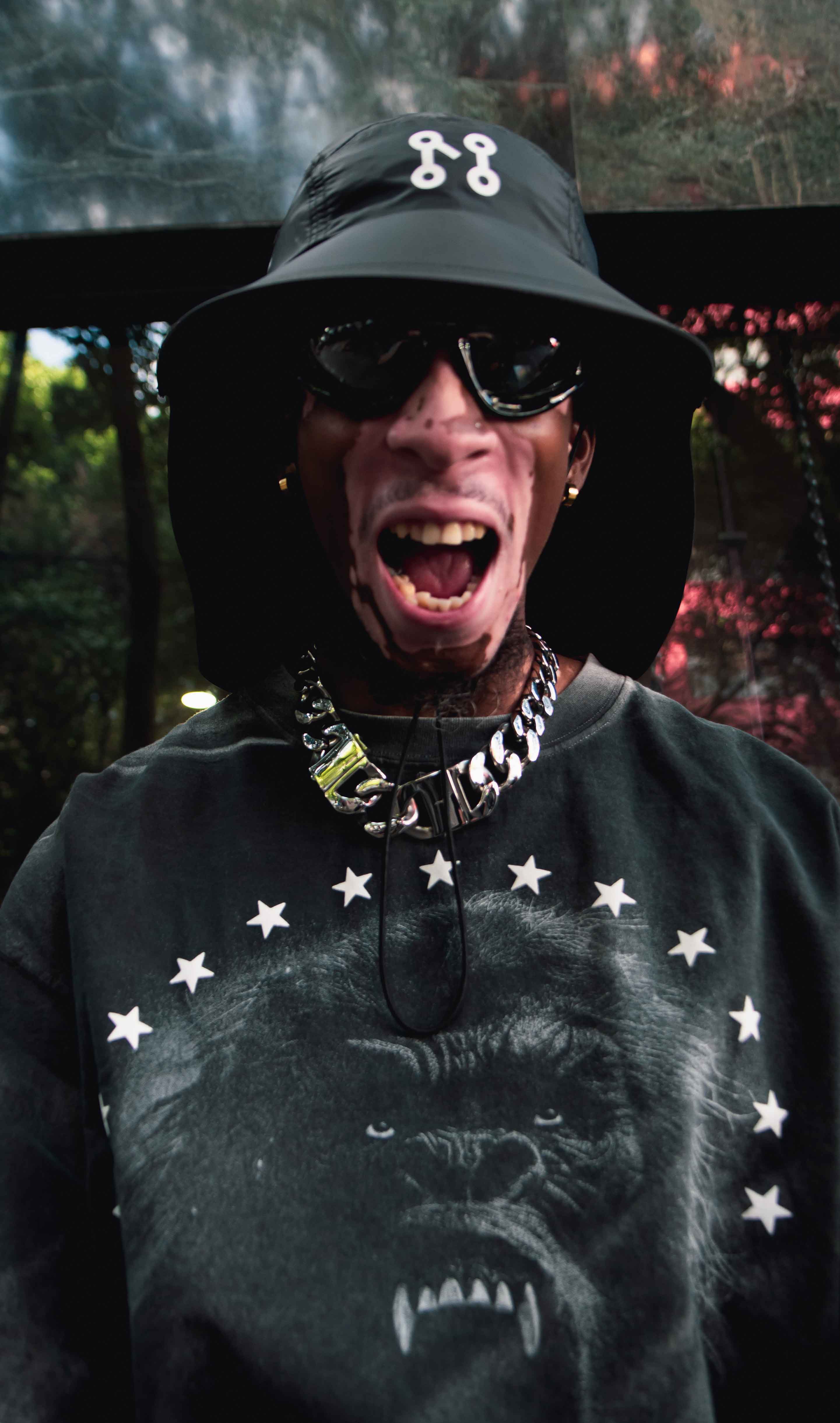 Wearing the El Protector bucket hat from Machina - Wearable Technology, along with stylish sunglasses and a graphic shirt featuring a gorilla design, the person radiates joy with an expressive open-mouthed smile. The background seamlessly blends wilderness vibes and Forest's Shadow, creating an intriguing mix of greenery and shadowy depths.