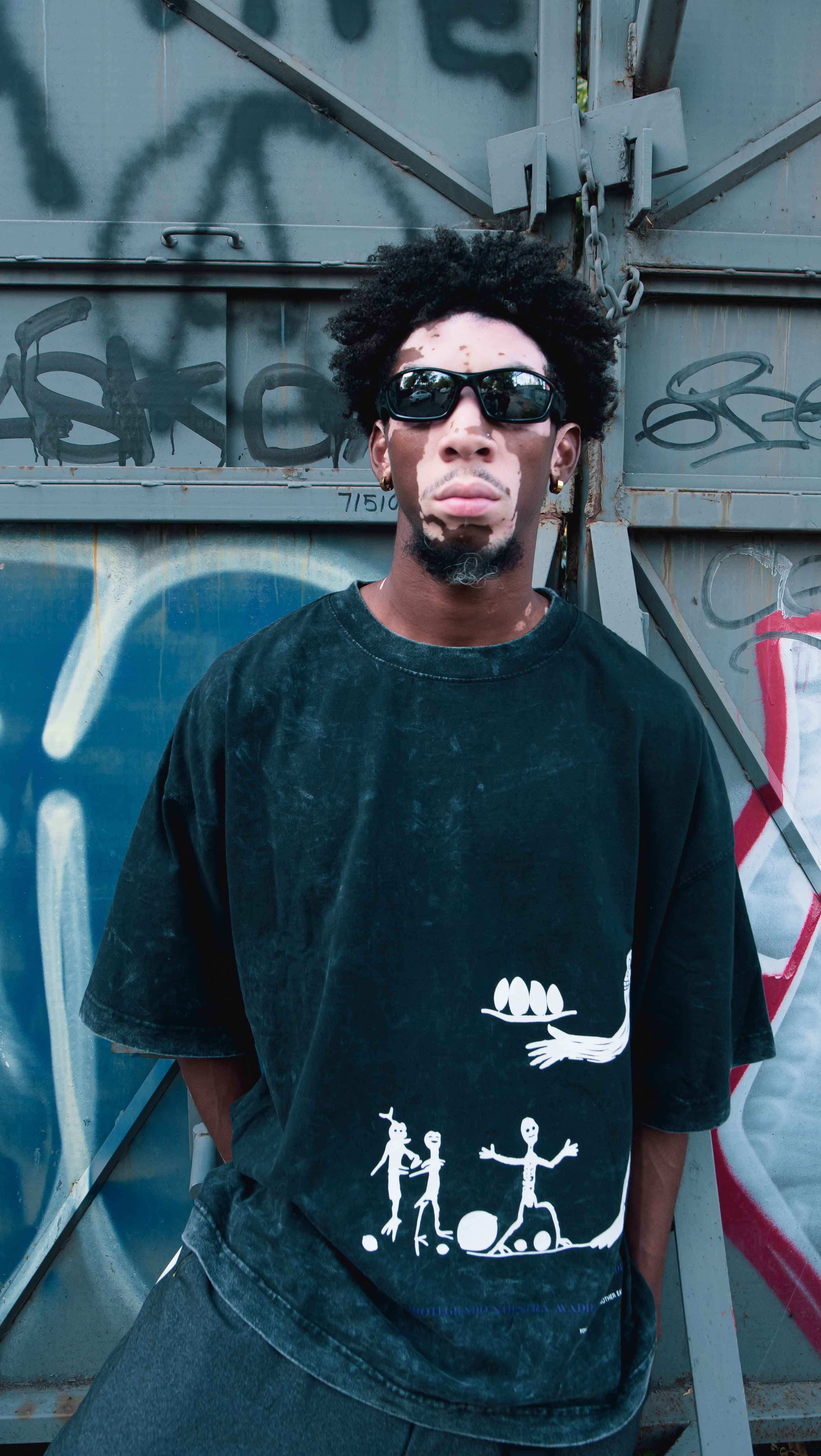 A model stands in front of a graffiti-covered wall, wearing a black Los Guardianes T-shirt by Machina - Wearable Technology, featuring cryptid artwork and sunglasses. Their voluminous and curly hairstyle complements the urban, artistic vibe of the setting.