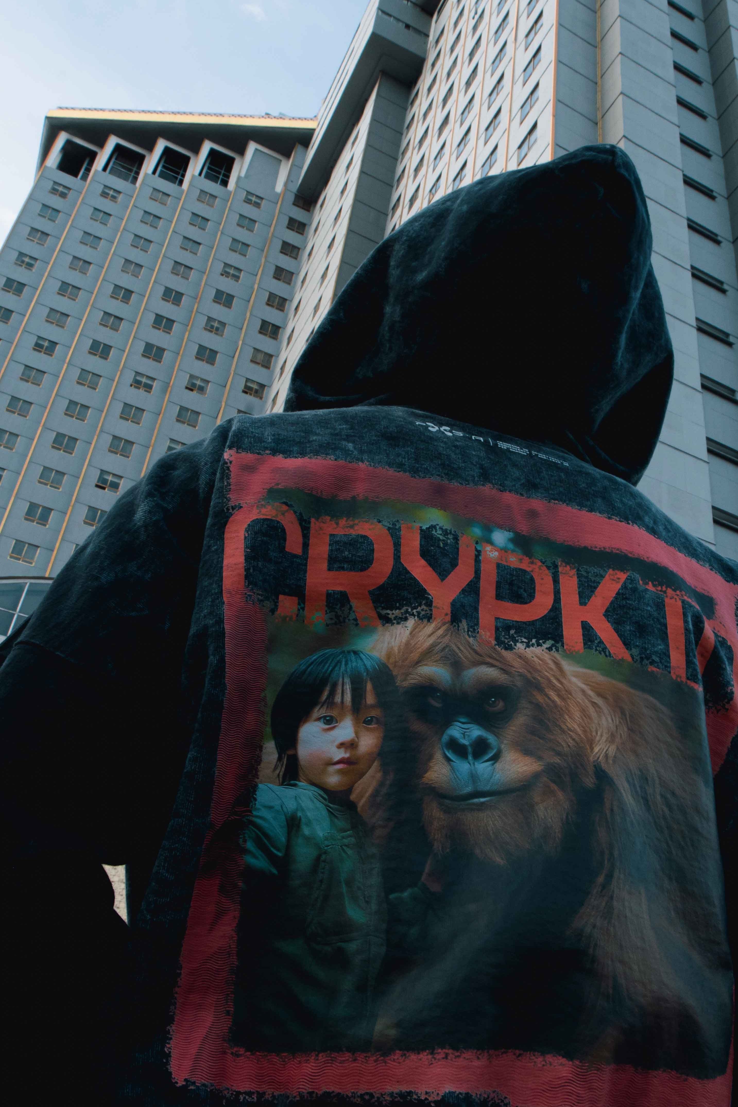 A person wearing a Crypkid hooded jacket, made from soft French terry cotton, stands with their back to the camera, facing tall buildings. The jacket displays a colorful graphic on the back featuring a child and an orangutan along with the words "CRYPK T," celebrating an unexpected friendship. This product is from Machina - Wearable Technology.