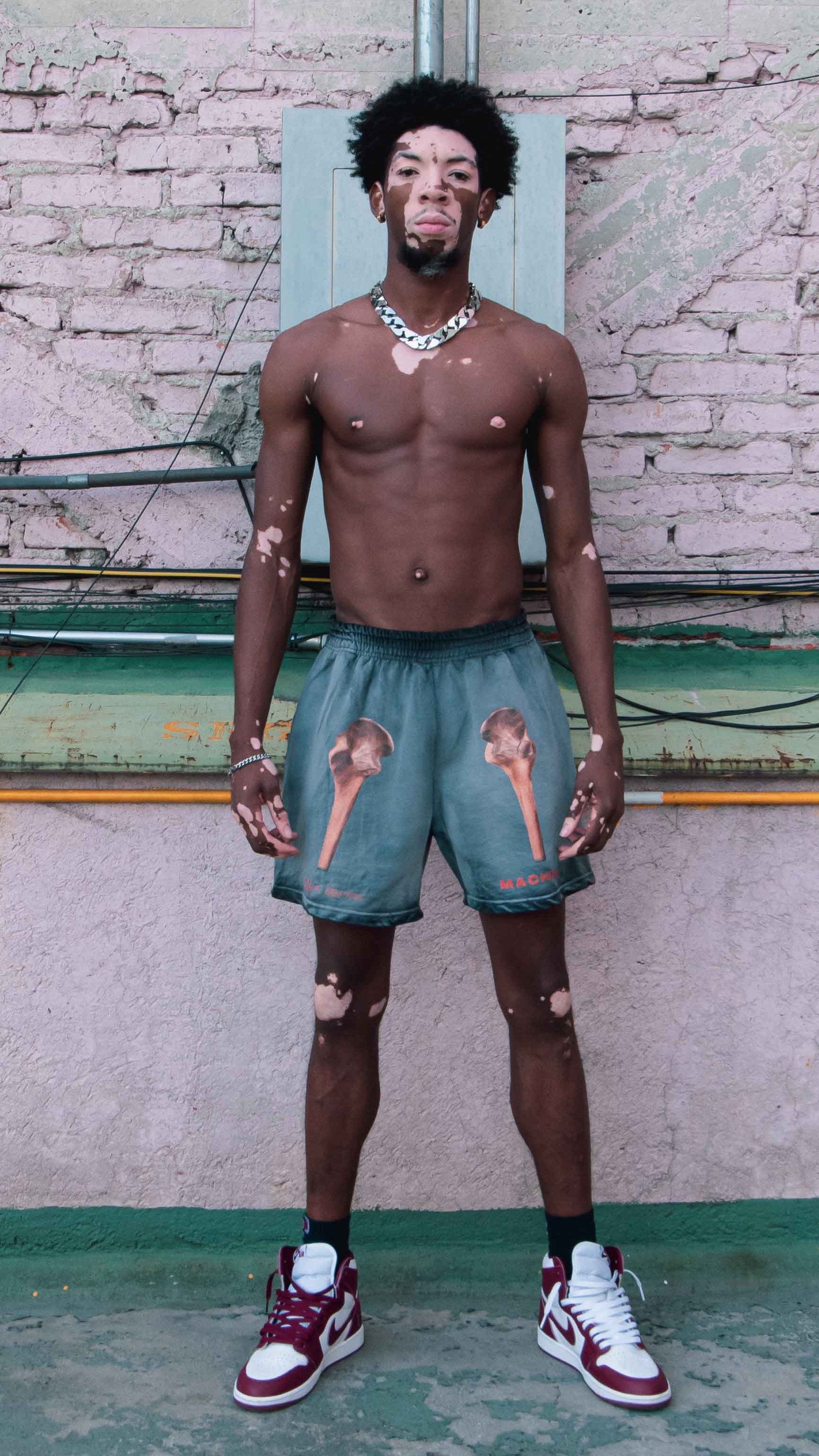 A model stands confidently against a textured brick wall. They are shirtless, wearing Machina - Wearable Technology's Pie Grande Skeleton Shorts, and sporting red and white sneakers. The individual is accessorized with a chain necklace, exuding a casual style.