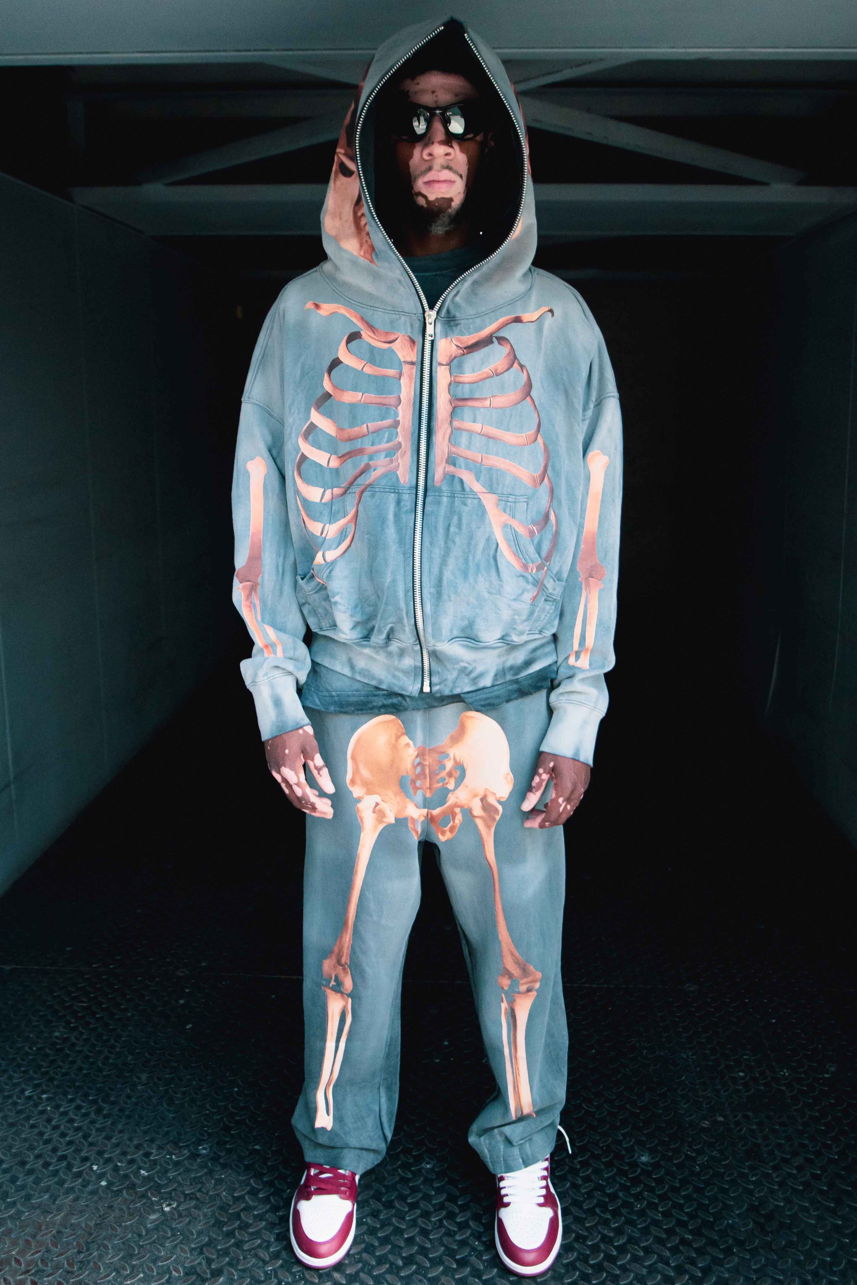 A mythical enthusiast stands in the shadows, embodying an air of mystery reminiscent of Bigfoot Skeleton myths. They wear the Pie Grande Skeleton set—a stylish ensemble by Machina - Wearable Technology— featuring a french terry cotton hoodie adorned with a ribcage design and pants displaying a pelvis motif, with hands resting by their sides.