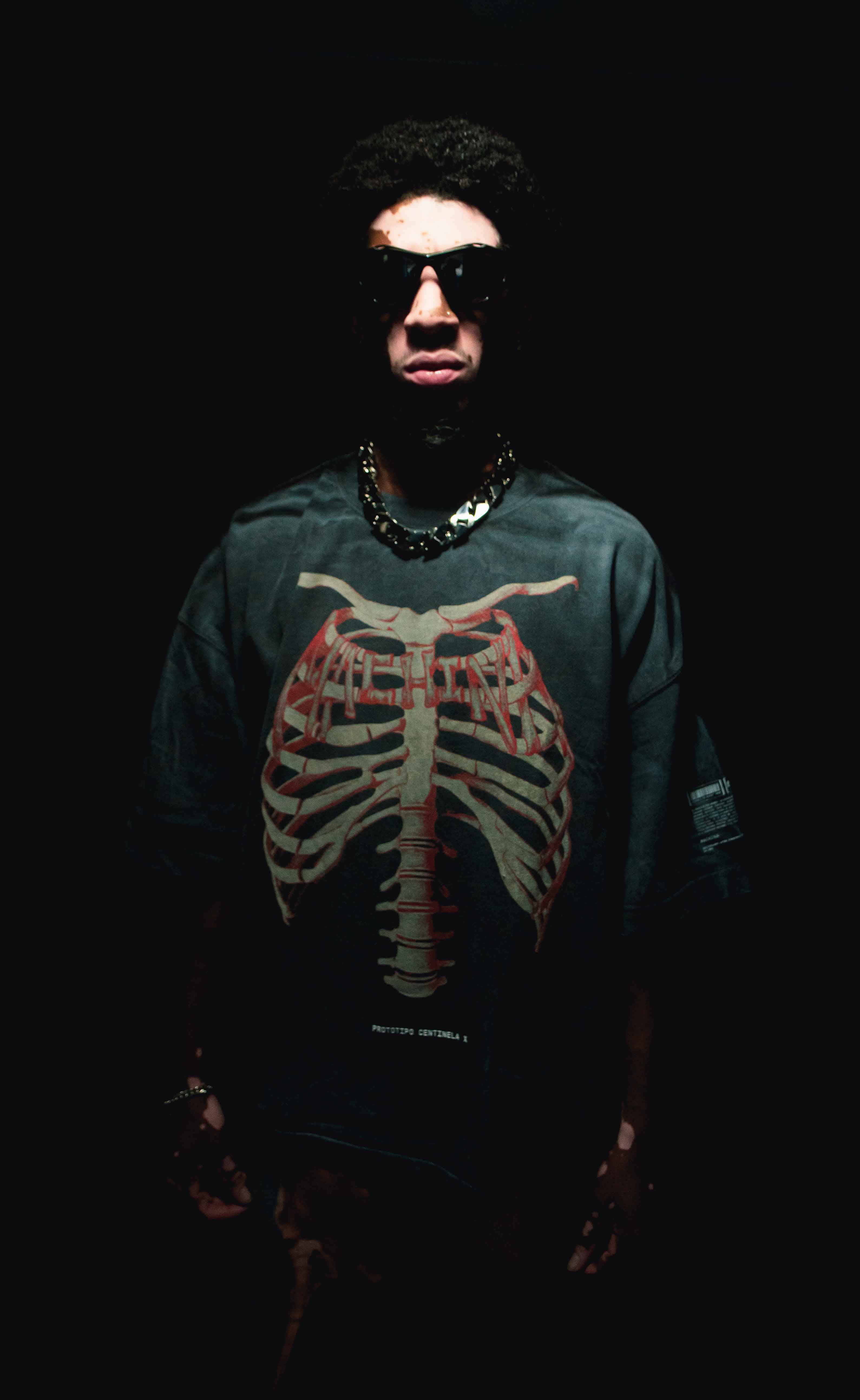 A person wearing sunglasses and a dark T-shirt featuring The Bigfoot Project's large skeleton ribcage design stands ominously against a black background, capturing the mysterious allure of government experiments. The lighting creates a dramatic, moody effect, enhancing the intrigue of Machina - Wearable Technology.