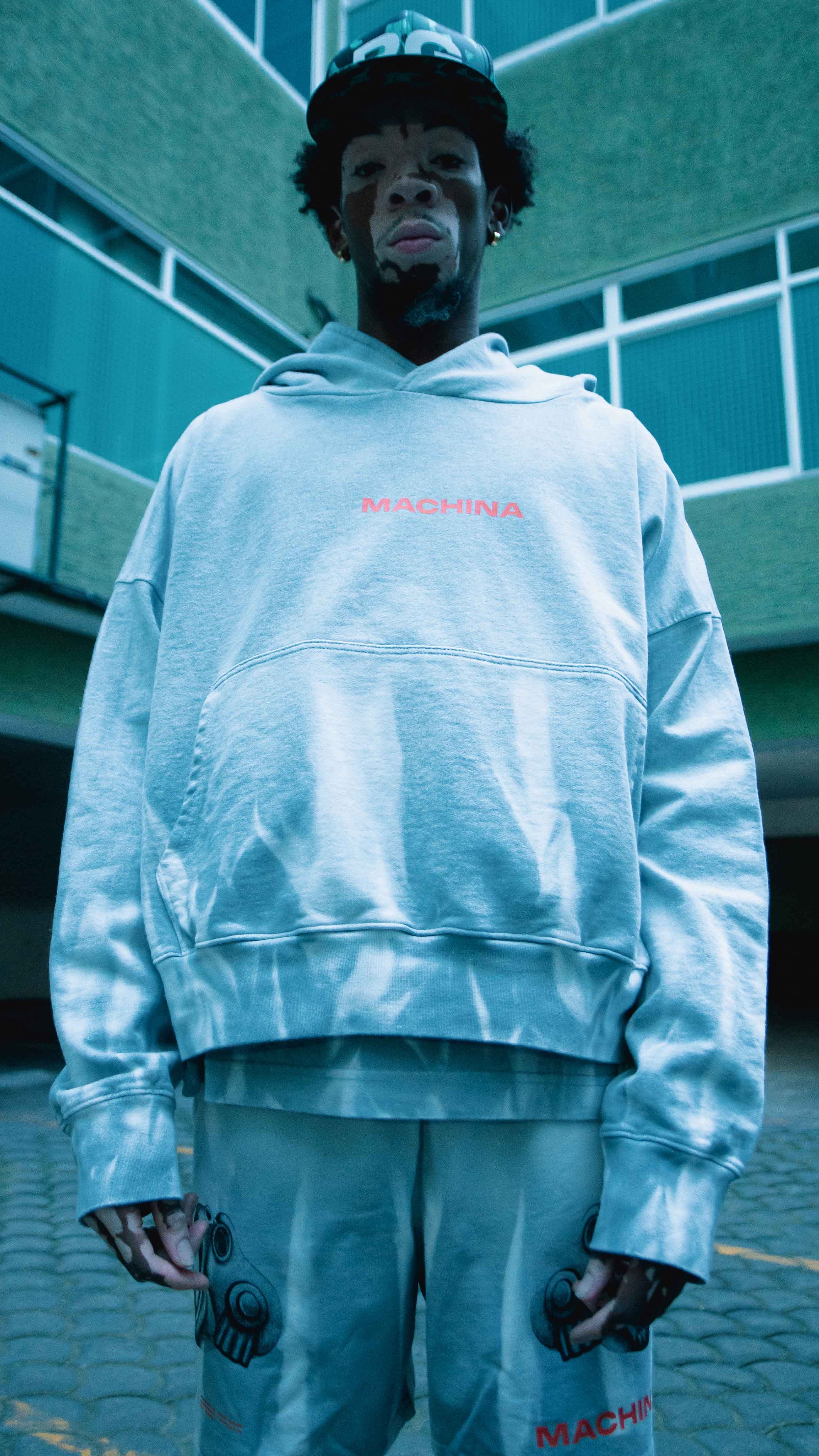 A person stands outdoors, wearing a loose-fitting Coatlicue Hoodie by Machina - Wearable Technology made of French terry cotton, along with matching shorts featuring the MACHINA logo in red. They adorn mask-like face paint and are positioned beneath a building with green-tinted windows, exuding the essence of an Aztec goddess.