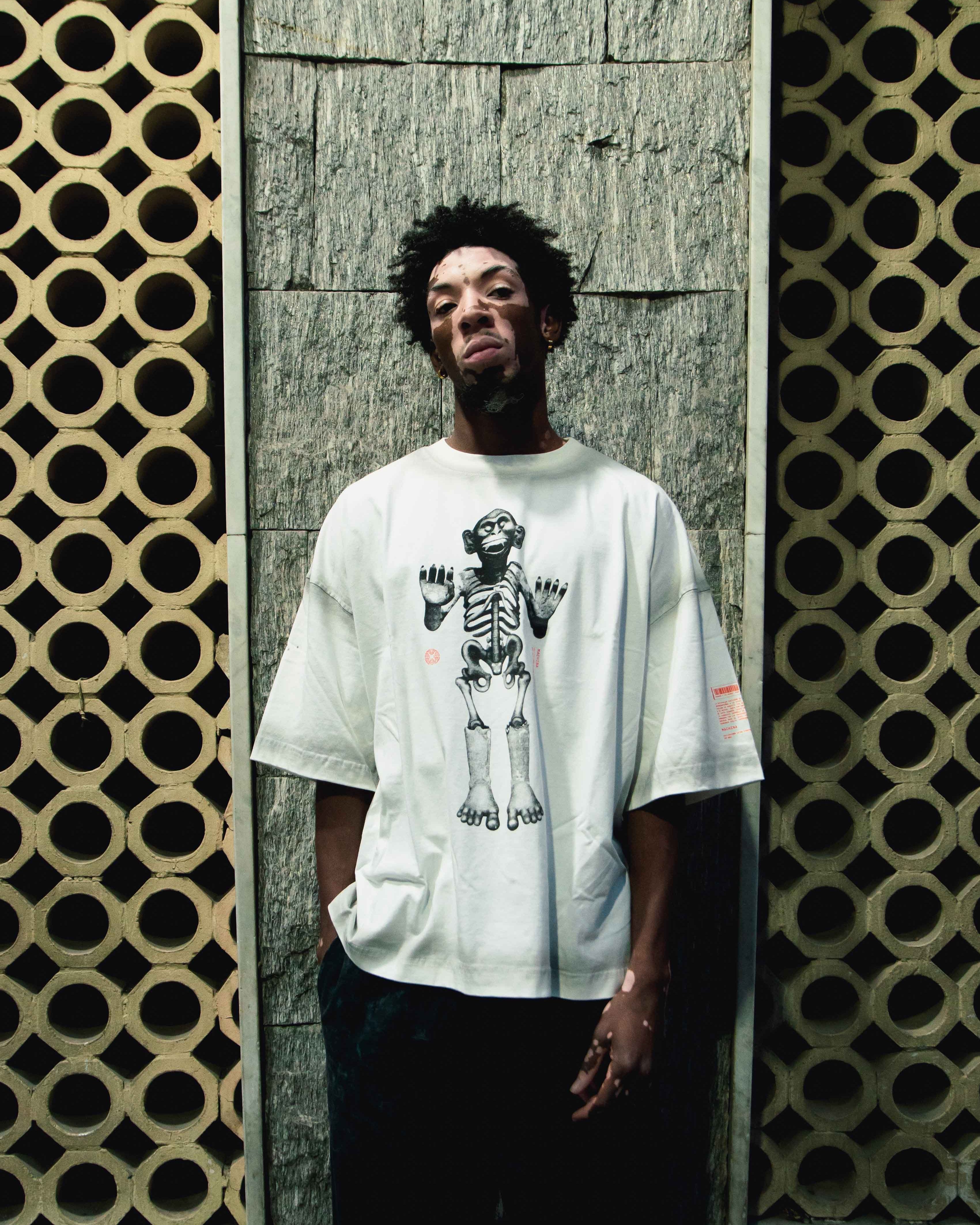 A person with short curly hair is leaning against a wall adorned with a geometric pattern reminiscent of Tenochtitlán. They are wearing a white Mictlantecuhtli t-shirt featuring a skeleton graphic from Machina - Wearable Technology. The background includes circular cutout designs on either side, creating a visually distinctive setting.