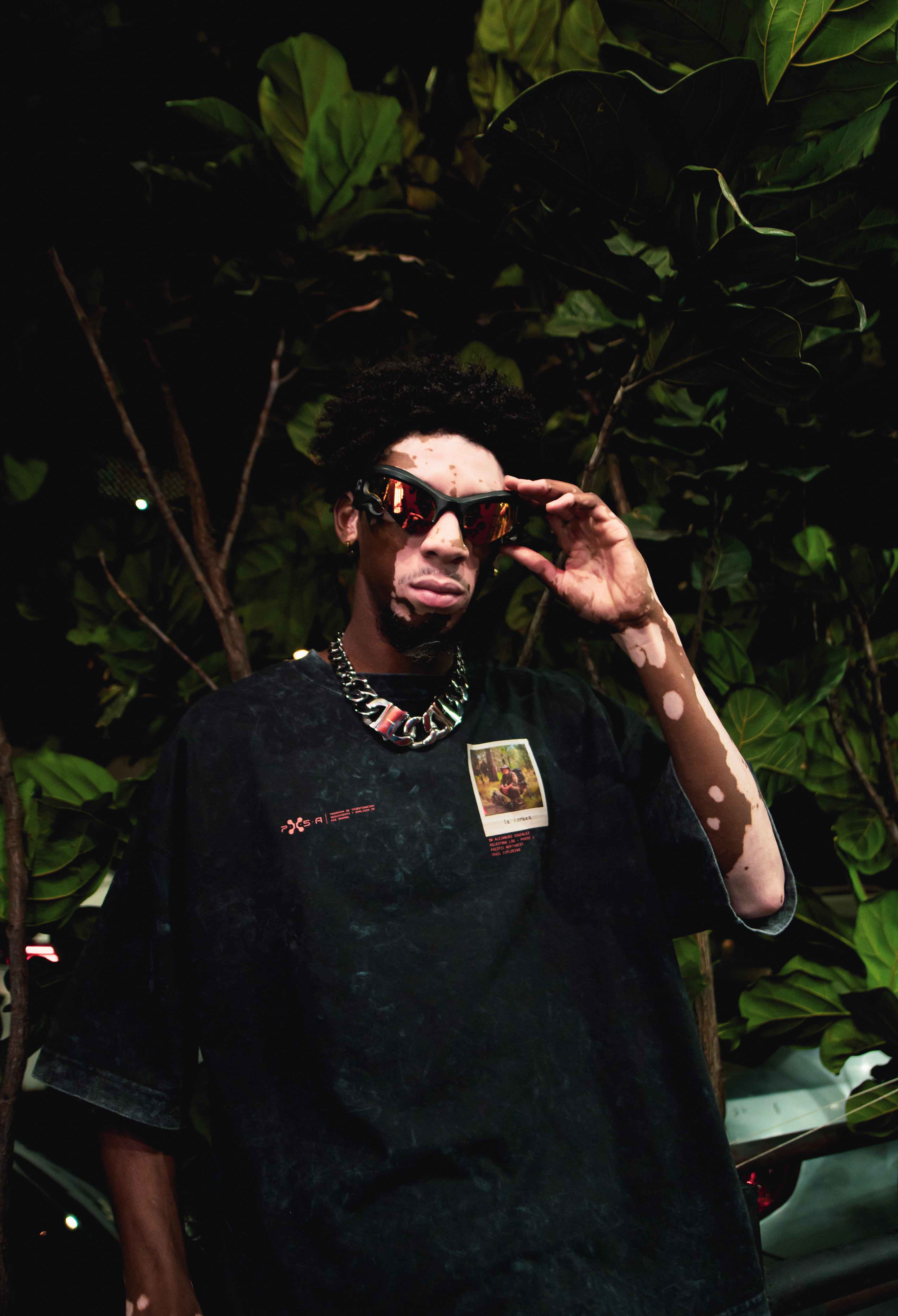 A person wearing Machina's La Sombra sunglasses and a black graphic T-shirt stands under leafy trees during a nighttime wilderness trek. They have their hands near the La Sombra sunglasses and wear a thick chain necklace. The setting is dimly lit, creating a moody atmosphere reminiscent of the Pacific Northwest.