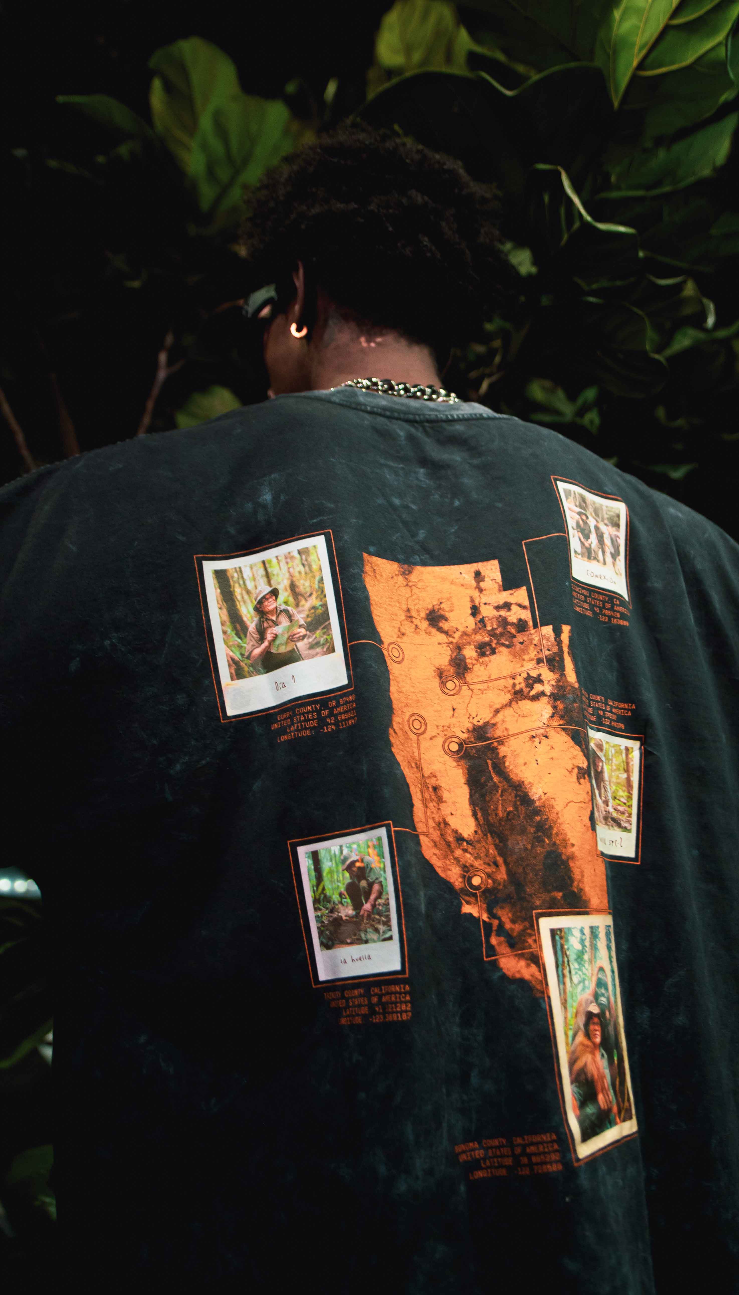 A person wearing the La Sombra T-shirt by Machina - Wearable Technology, which features an orange map and several small images printed on the back, stands amidst lush green foliage, appearing prepared for a Pacific Northwest wilderness trek.