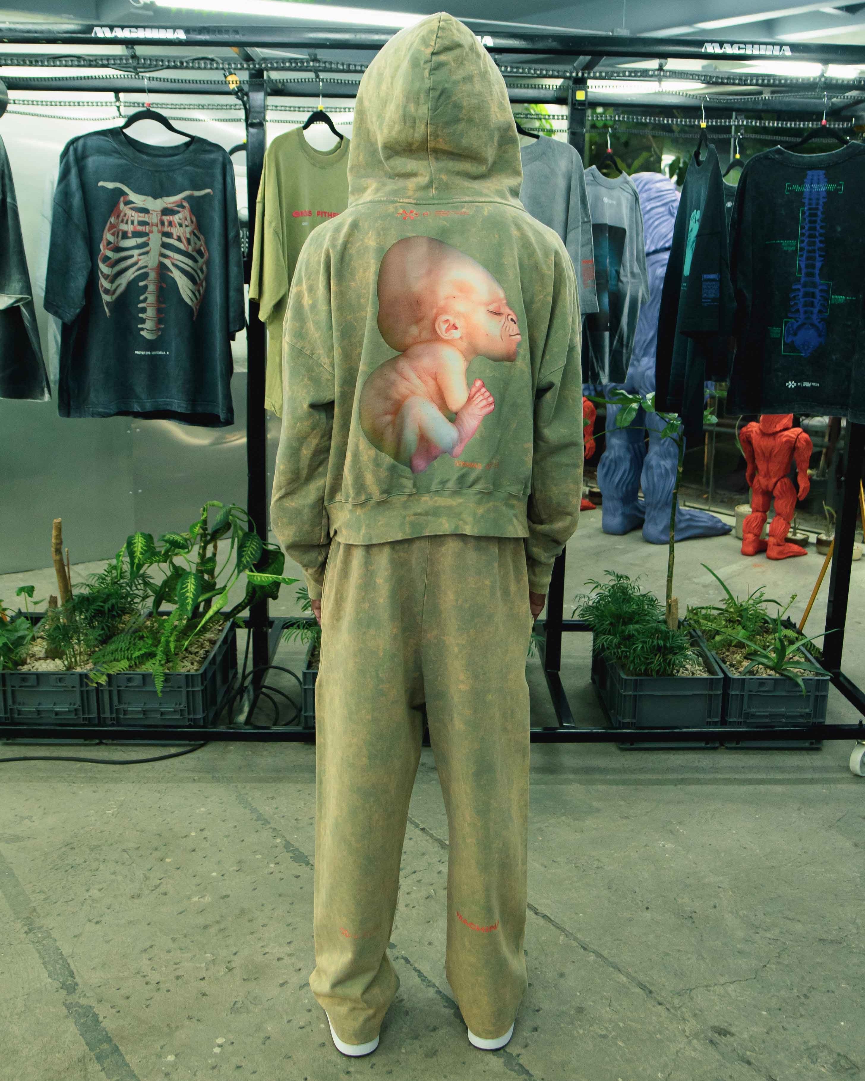 A person wearing a matching hoodie and pants, crafted from heavyweight French terry cotton adorned with a large Pie Grande Embryo print on the back, stands in a clothing store. The space is filled with artistic shirts and lush plants, exuding an inviting vibe, all under the brand Machina - Wearable Technology.