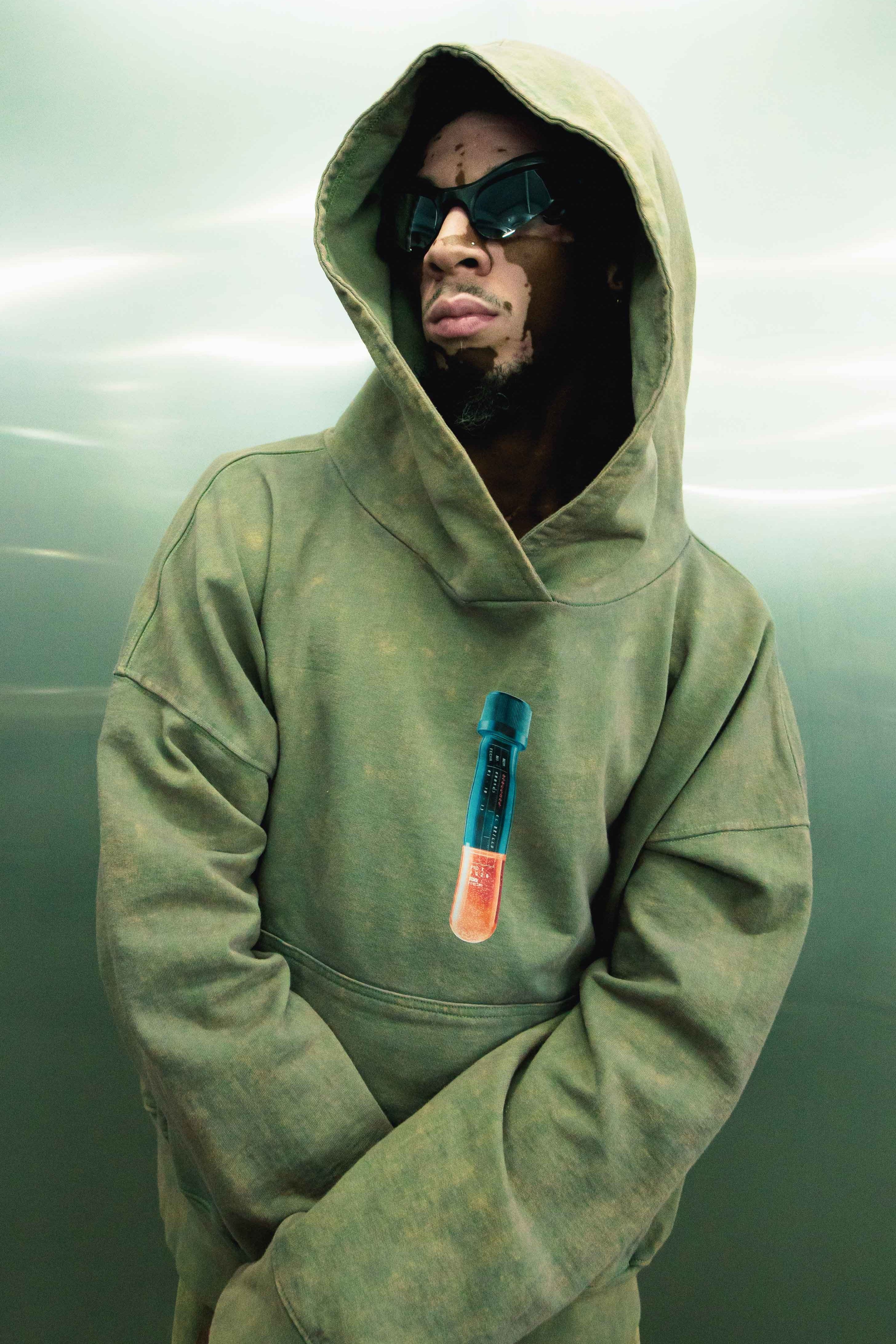 A mysterious figure wearing an oversized La Célula hoodie from Machina - Wearable Technology, with the hood up and dark sunglasses on, stands against a plain background. The hoodie features a vibrant test tube graphic on the front.