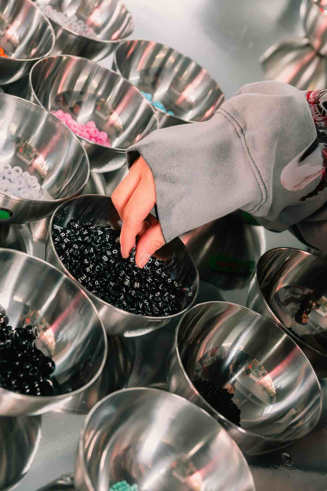 At the OUROBOROS DIY CHARM WORKSHOP by Machina Wearable Technology, a participant in a gray sleeve picks black alphabet beads from a metal bowl surrounded by other colorful beads for custom jewelry creation.