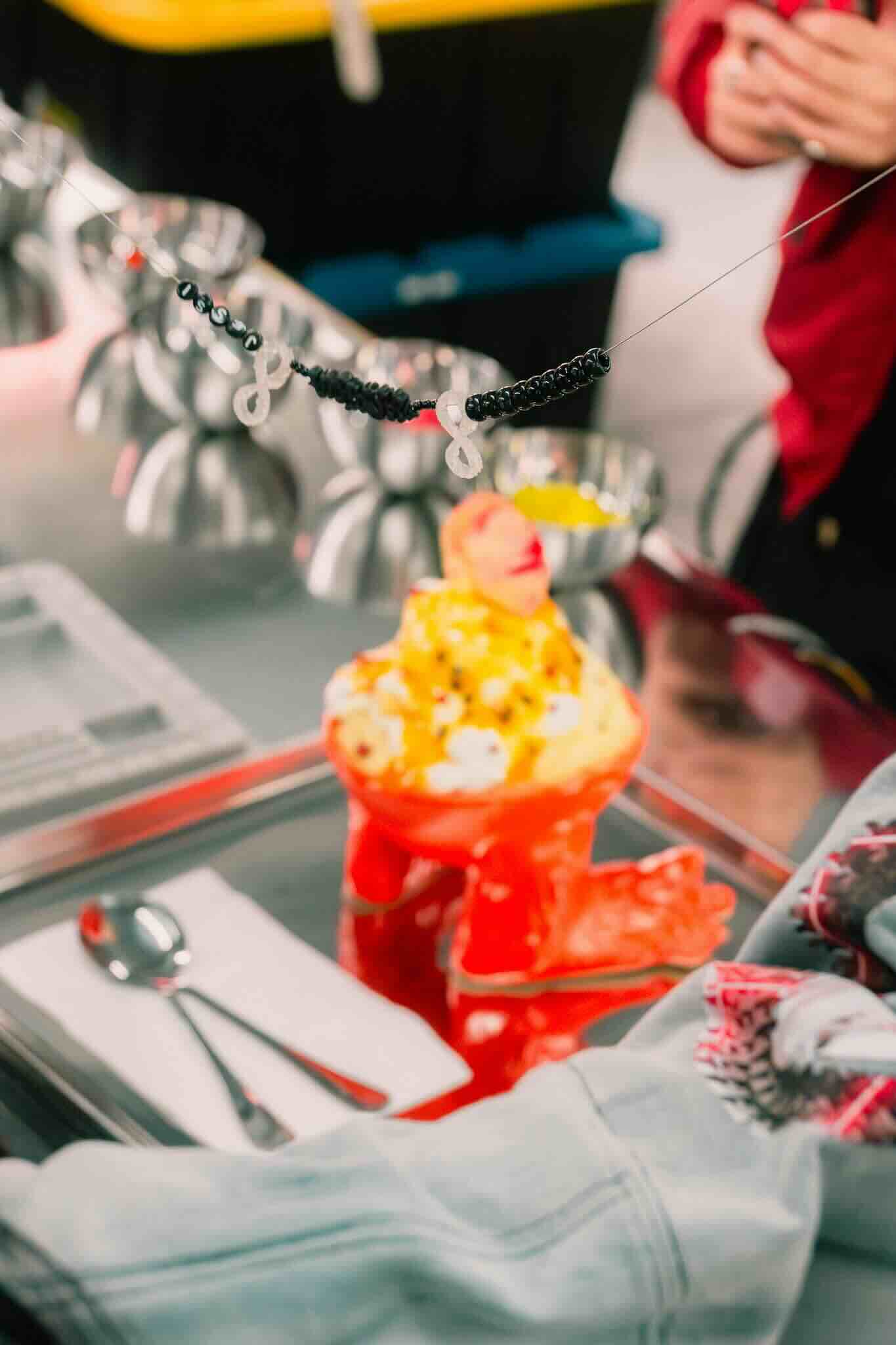 A vibrant dessert in a red bowl sits on a tray with utensils and napkin. In front, a custom beaded string from the OUROBOROS DIY CHARM WORKSHOP by Machina Wearable Technology hangs above the treat.