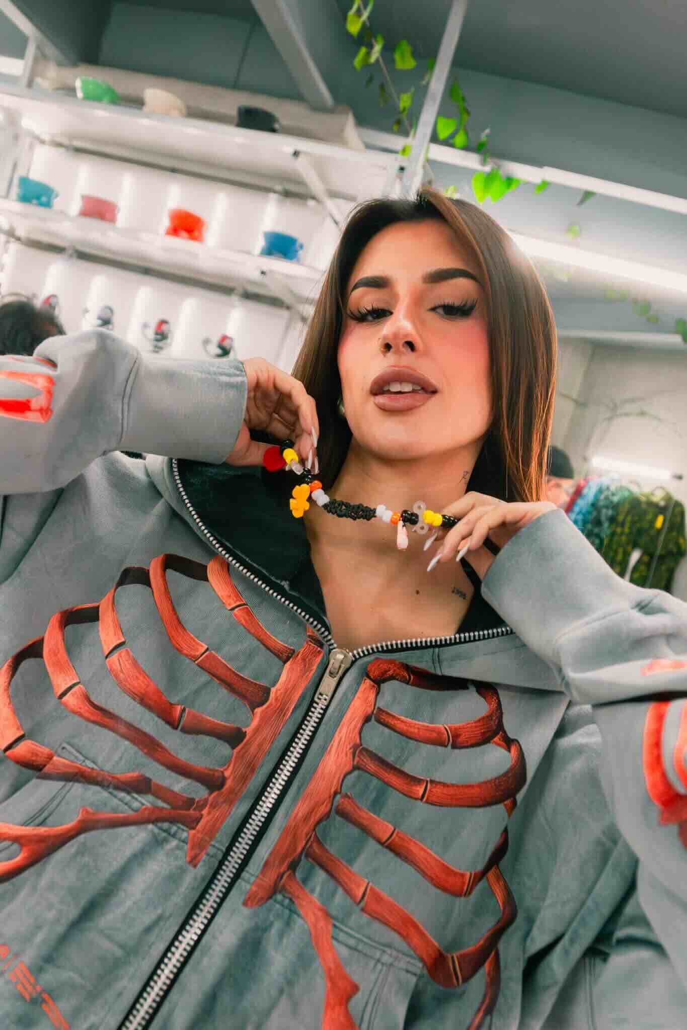 A woman indoors holds a colorful beaded necklace from the OUROBOROS DIY CHARM WORKSHOP by Machina Wearable Technology. She wears a gray jacket with a red ribcage design; shelves with jars and plants are in the background.