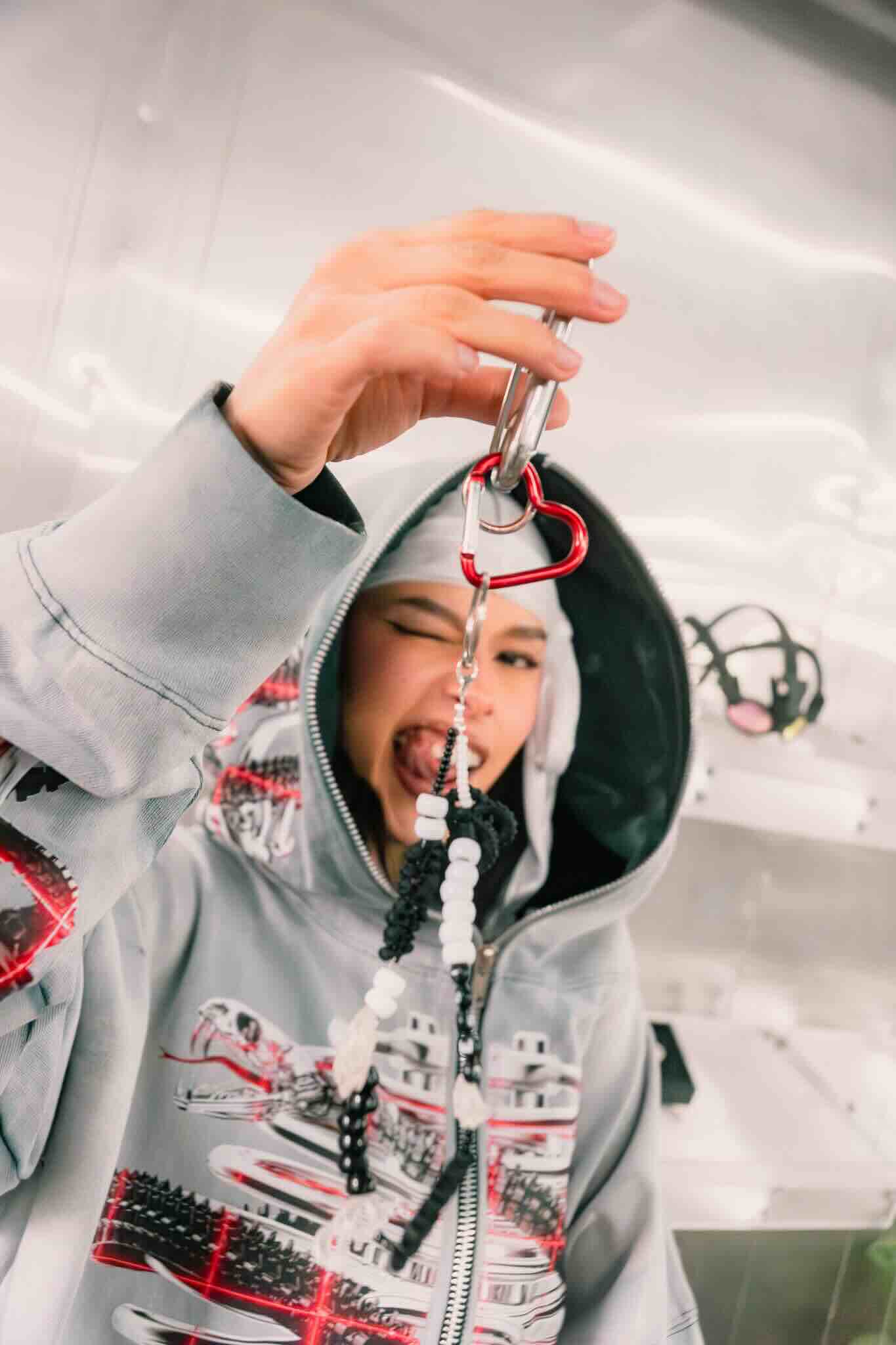 A person in a gray hoodie winks and shows off a beaded keychain with a red carabiner—get inspired at the OUROBOROS DIY CHARM WORKSHOP by Machina Wearable Technology and create your own custom jewelry in a bright, modern space!.