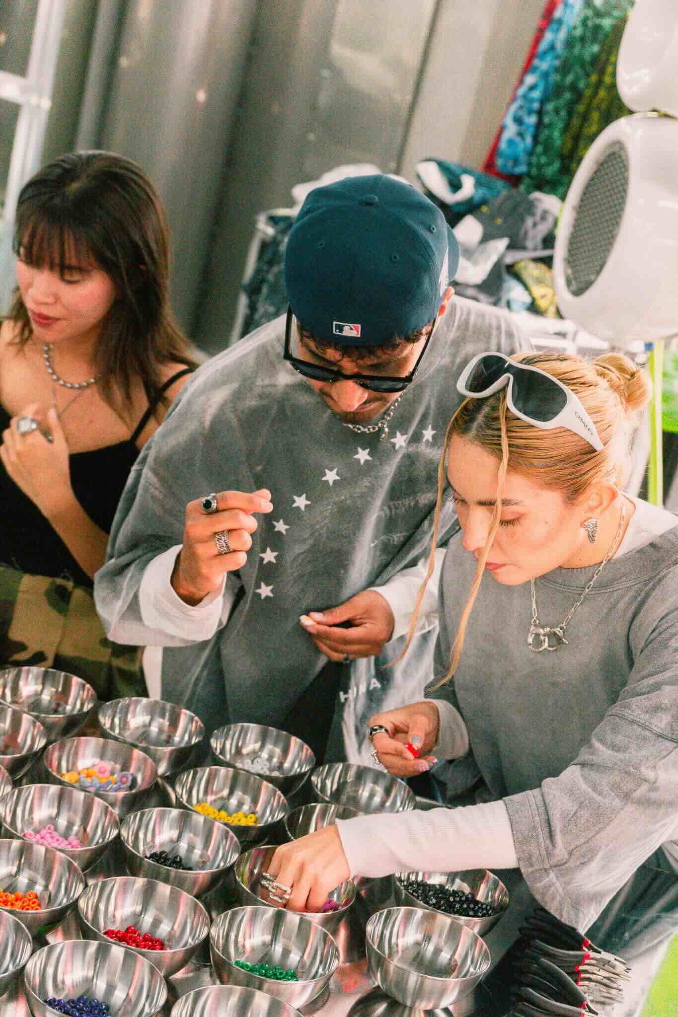 At the OUROBOROS DIY CHARM WORKSHOP by Machina Wearable Technology, three people gather around a table of vibrant beads and metal bowls, selecting pieces and possibly designing custom jewelry in a fun, creative setting.