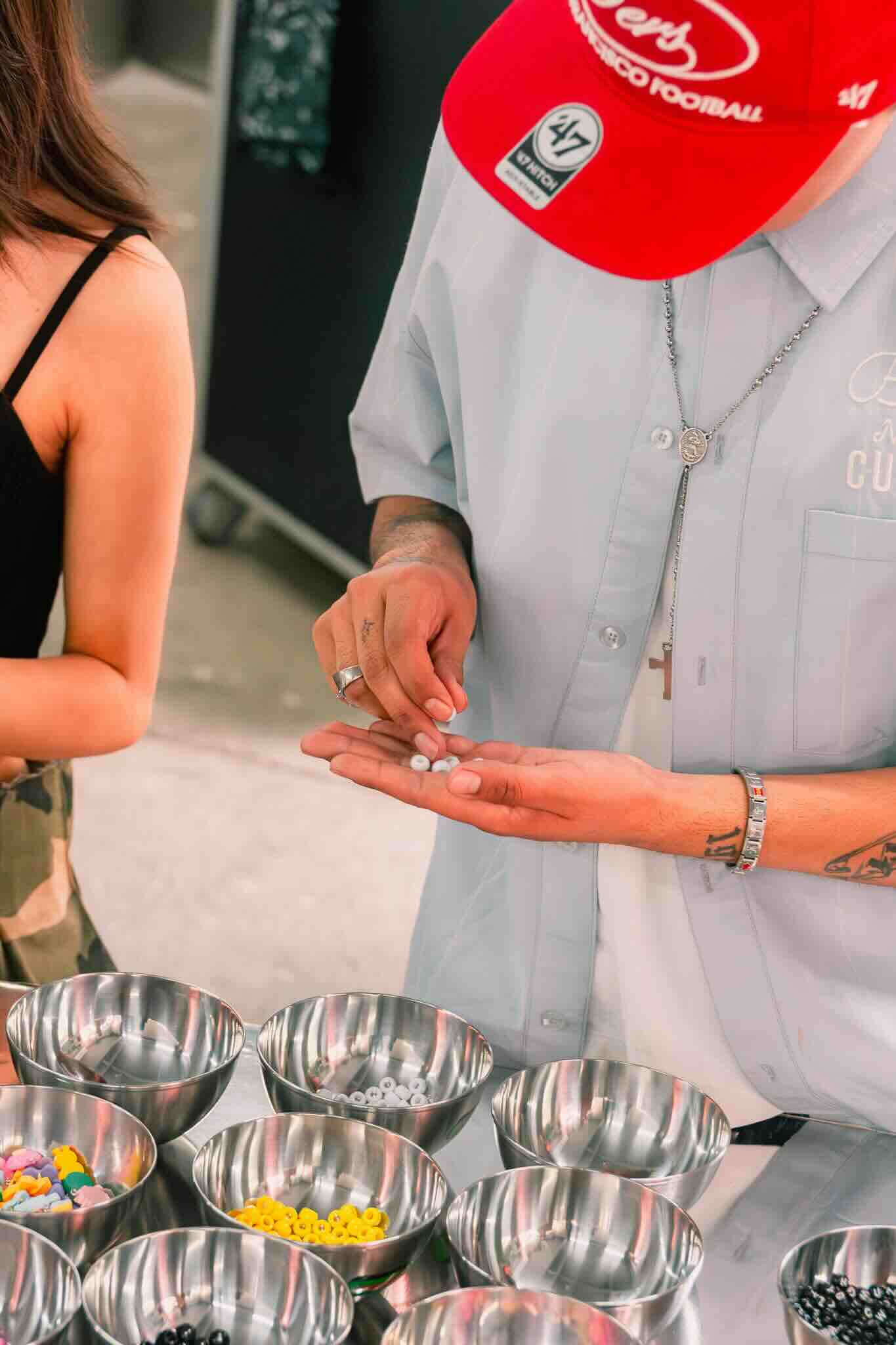 At the OUROBOROS DIY CHARM WORKSHOP by Machina Wearable Technology, a participant in a red cap selects colorful beads and resin pieces from metal bowls, while another attendee enjoys this creative, hands-on experience.