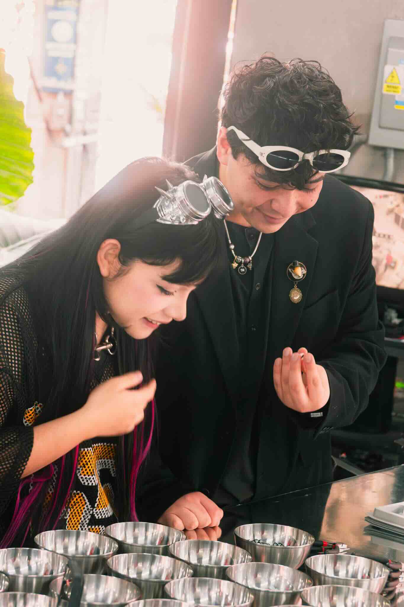 Two people wearing black clothing and goggles smile as they create custom jewelry at the OUROBOROS DIY CHARM WORKSHOP by Machina Wearable Technology, looking excited and engaged while working with metal bowls on a table.