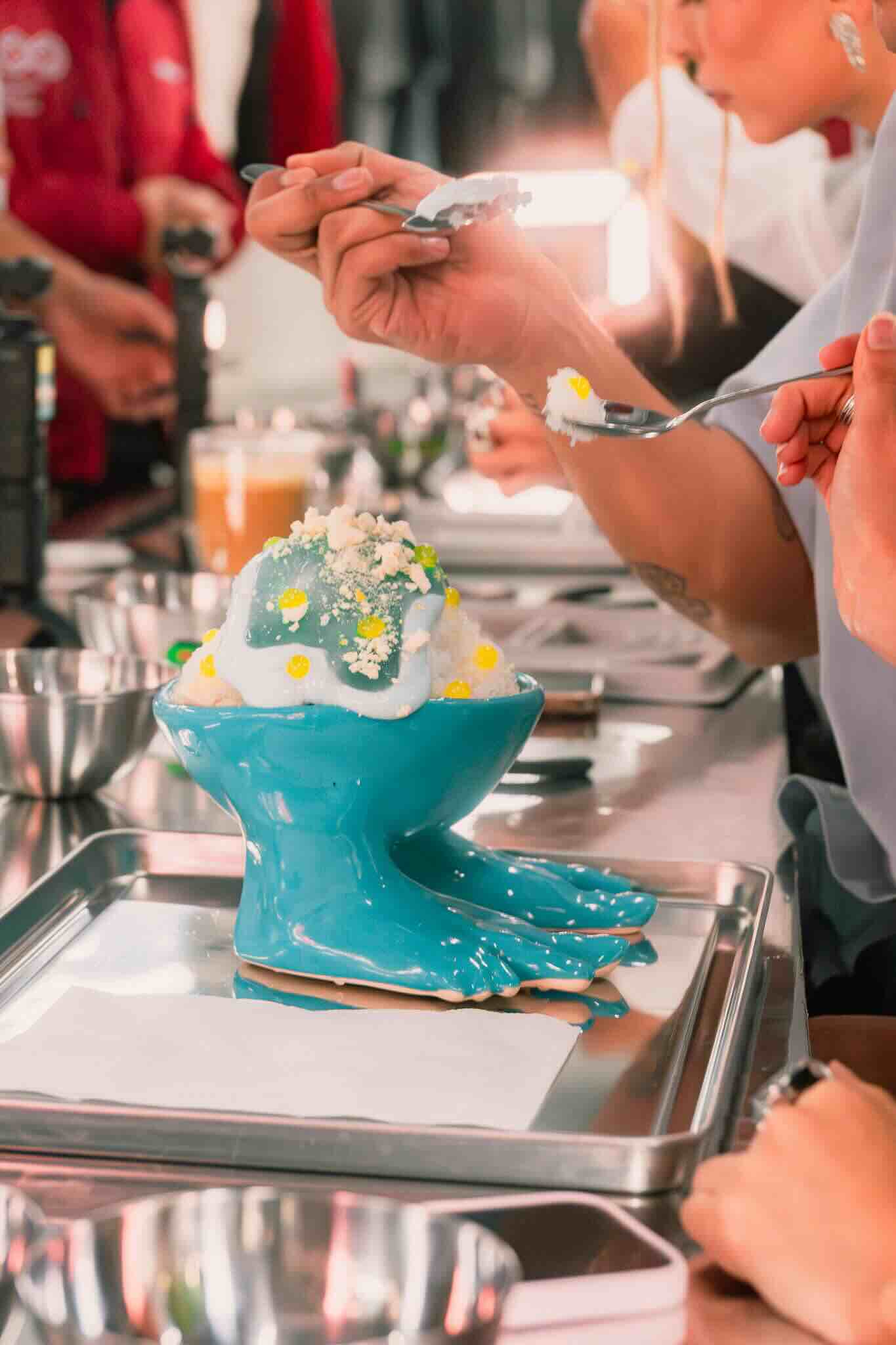 At a kitchen counter set up for the Machina Wearable Technology OUROBOROS DIY CHARM WORKSHOP, people decorate desserts with white cream and yellow accents in blue, foot-shaped bowls among trays and utensils.