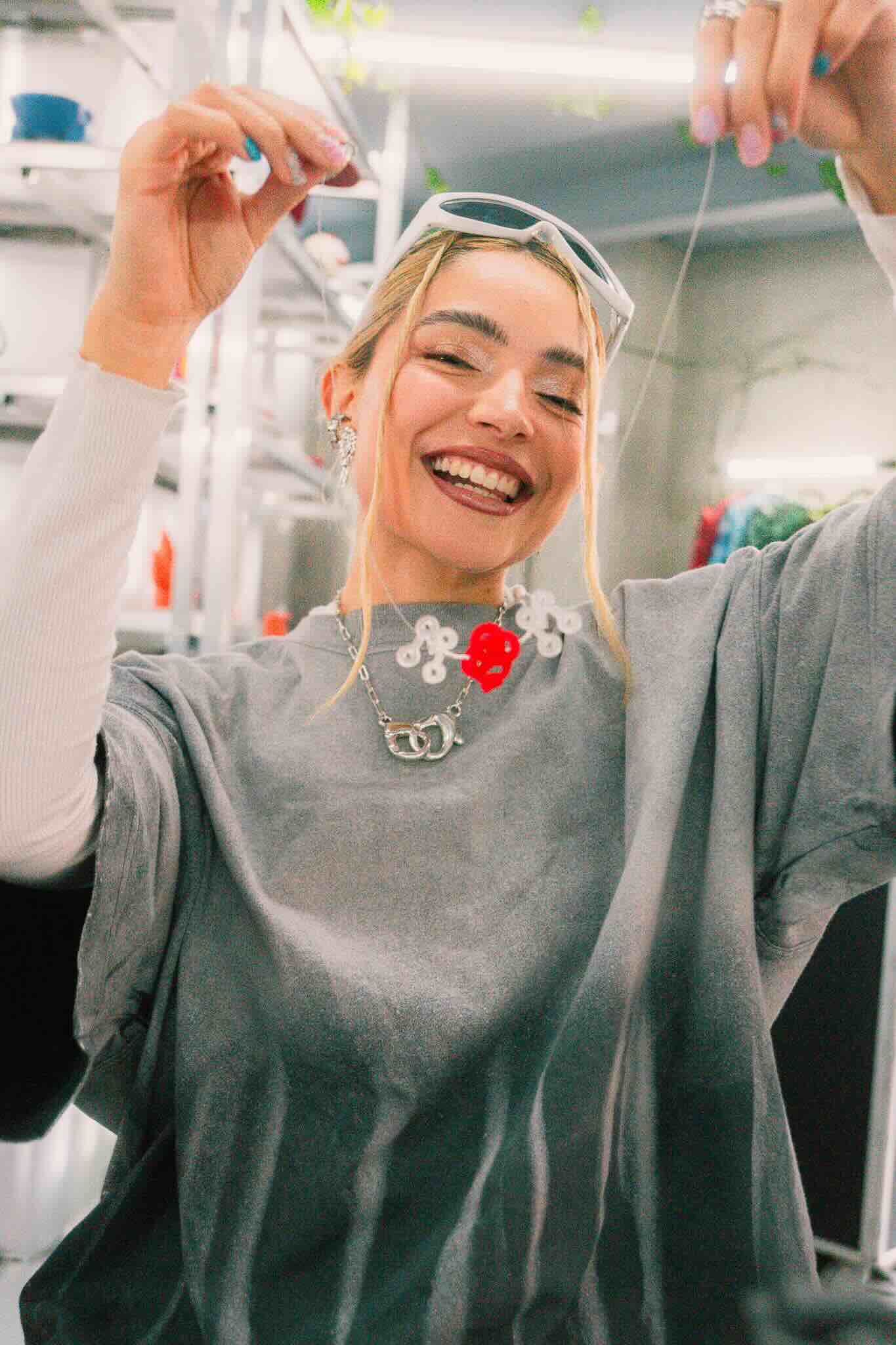 A smiling blonde woman wearing sunglasses and an oversized grey shirt holds up beaded creations at the OUROBOROS DIY CHARM WORKSHOP by Machina Wearable Technology in a bright indoor jewelry workshop.