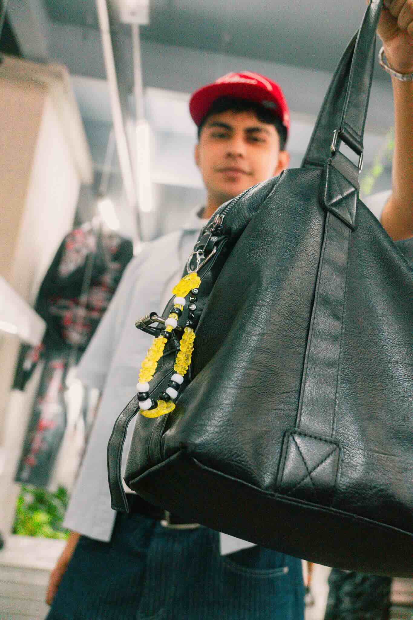 At the OUROBOROS DIY CHARM WORKSHOP by Machina Wearable Technology, a person in a red cap and light shirt holds a black leather bag with a yellow and black beaded keychain, surrounded by shelves and hanging items.