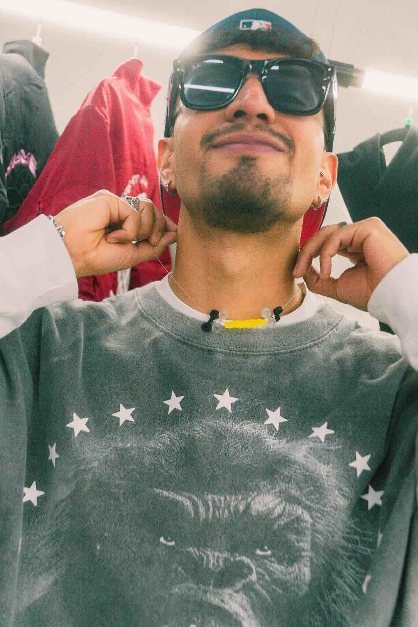 A man in sunglasses, a backward cap, and gorilla-print sweatshirt smiles as jackets hang on a rack. Nearby, Machina Wearable Technology's OUROBOROS DIY CHARM WORKSHOP resin pieces are displayed, creating a casual, candid atmosphere.