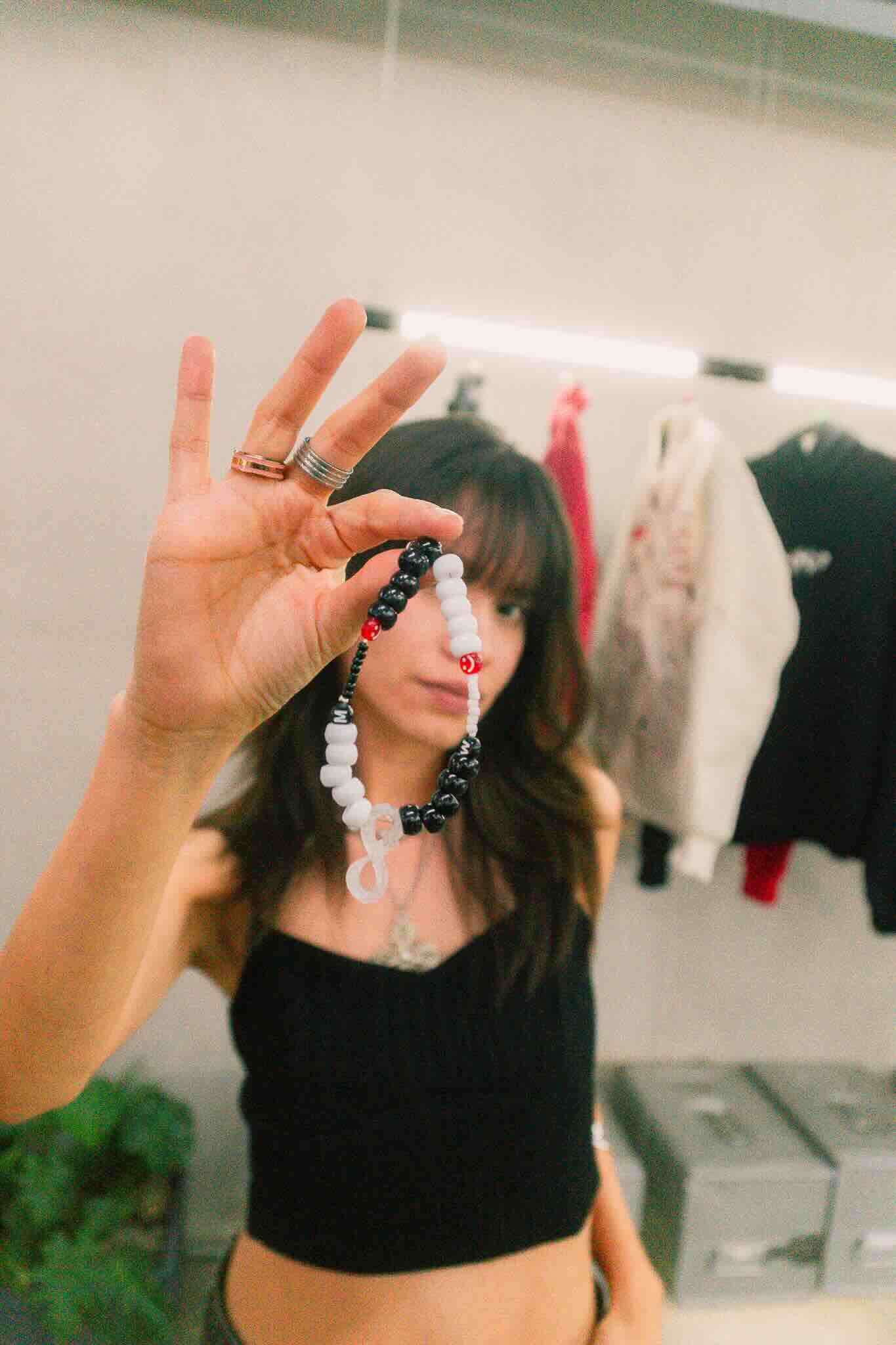 A woman in a black top holds up a Machina Wearable Technology OUROBOROS DIY CHARM WORKSHOP bracelet with black, white, and red beads, partially covering her face as clothing hangs on a blurred rack in the background.