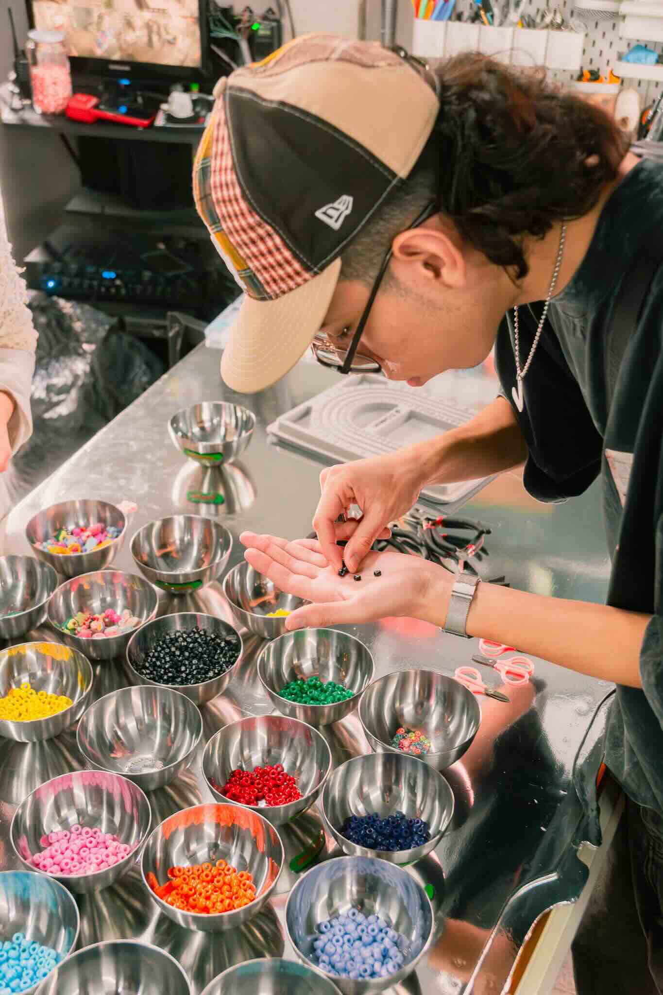 In a bright, organized workspace, hands focus on sorting colorful beads and resin for the OUROBOROS DIY CHARM WORKSHOP by Machina Wearable Technology, with metal bowls reflecting their creative jewelry process.