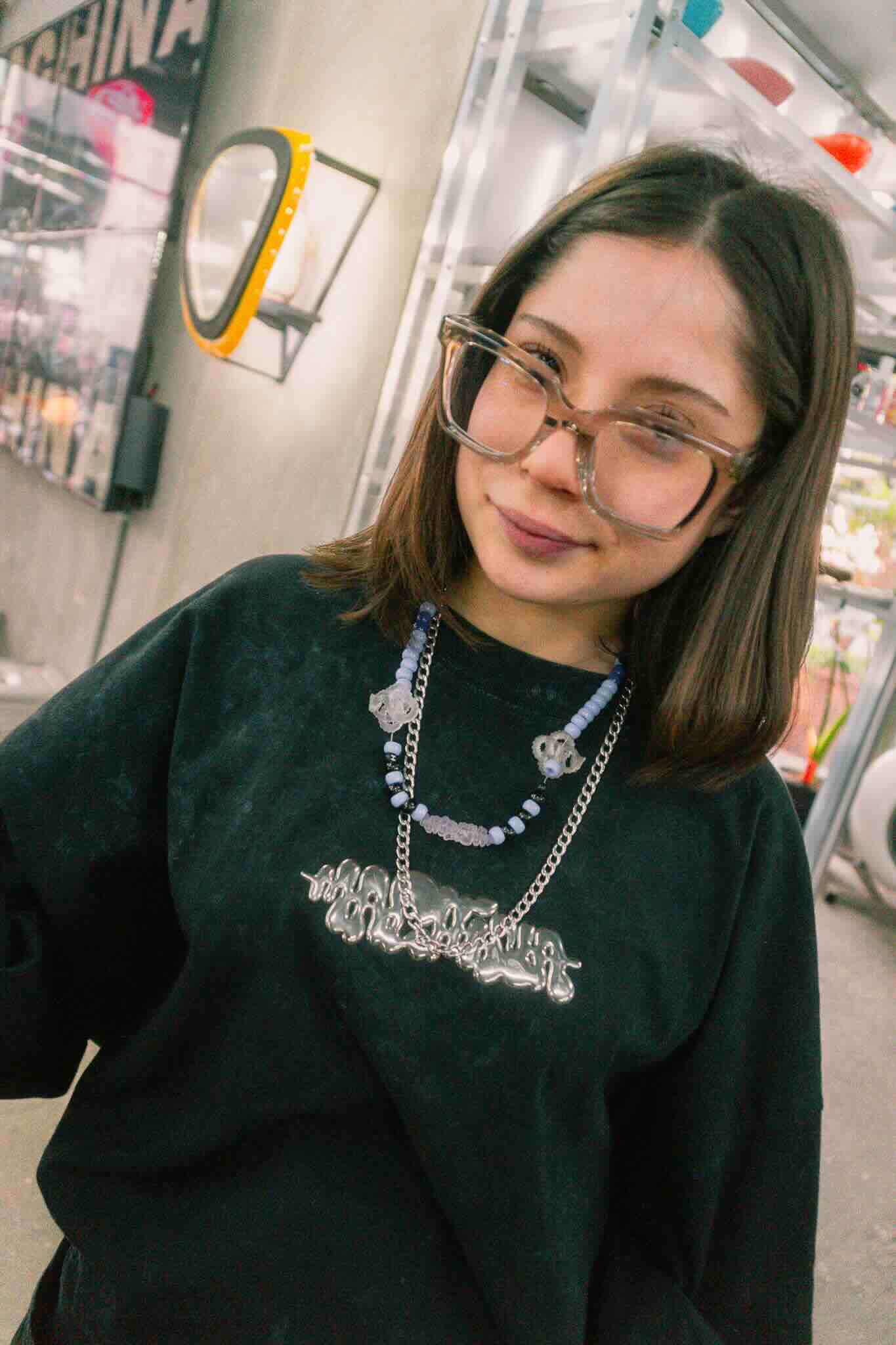 A young woman with straight brown hair wears oversized glasses and a black sweatshirt at the OUROBOROS DIY CHARM WORKSHOP by Machina Wearable Technology, smiling softly in a jewelry studio with shelves and a mirror behind her.