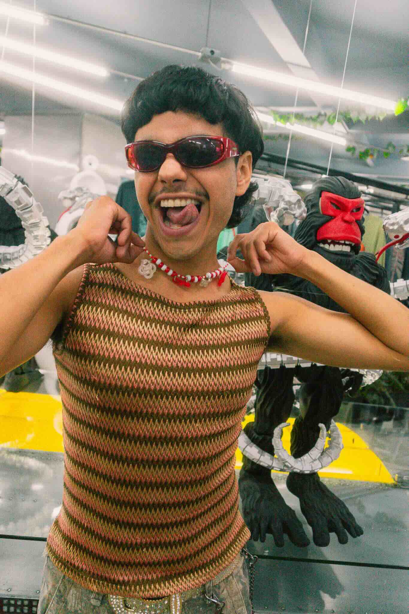 A person wearing Machina Wearable Technology’s OUROBOROS DIY CHARM WORKSHOP jewelry poses playfully in red sunglasses and a brown zigzag-patterned sleeveless top, with black gorilla statues with red faces and white chains behind them.