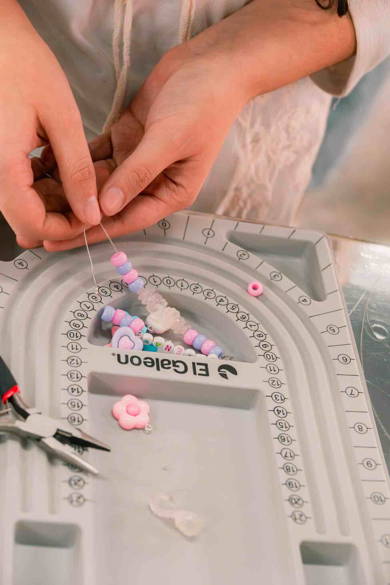 At the OUROBOROS DIY CHARM WORKSHOP by Machina Wearable Technology, someone crafts custom jewelry, stringing colorful beads and flower-shaped resin charms onto clear thread with a bead board—pliers are also on the table.