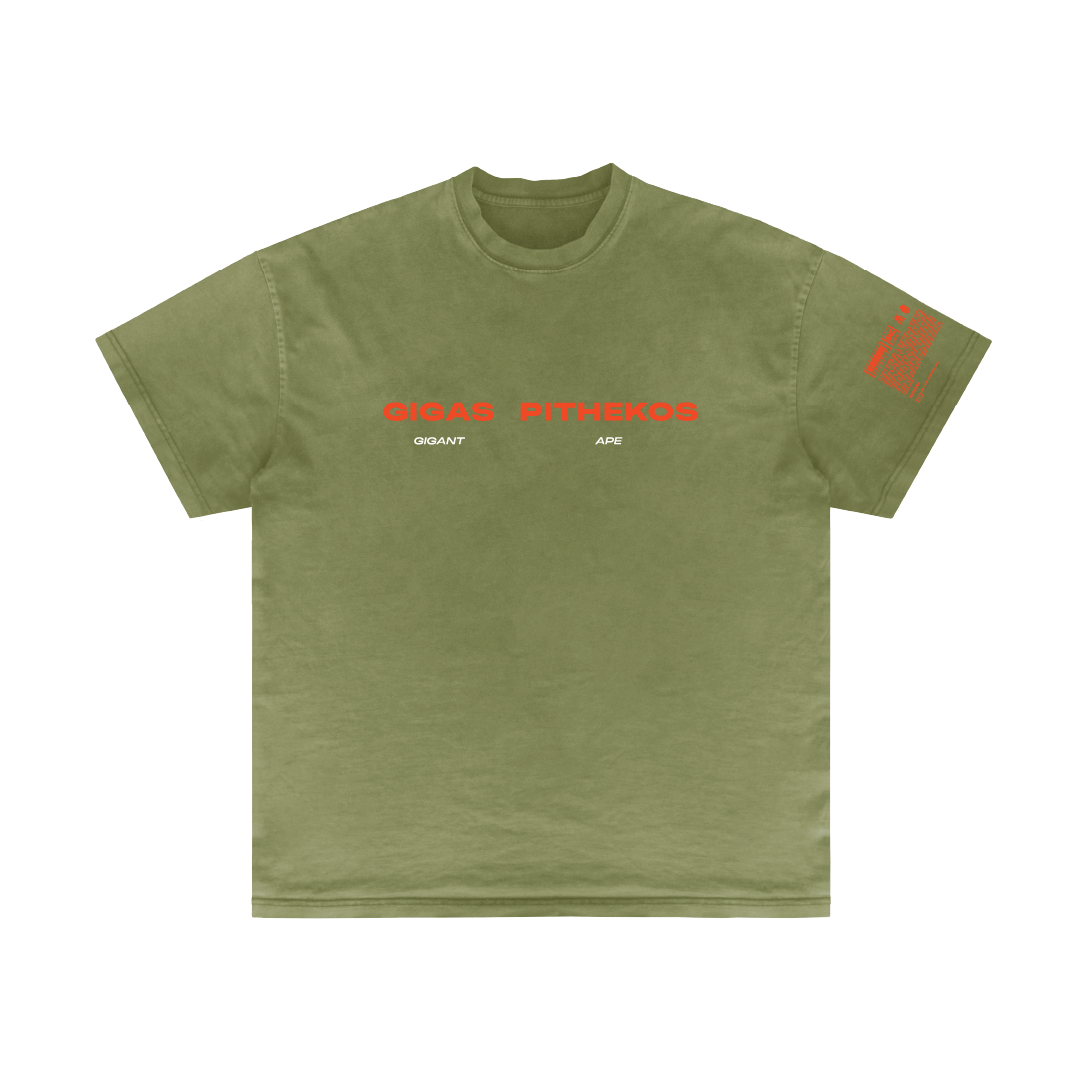 The olive green T-shirt from Machina - Wearable Technology, named "Gigas Pithekos," features bright red text across the chest displaying "GIGAS PITHEKOS," subtly referencing Bigfoot lore, with a smaller "GIGANT APE" underneath. The right sleeve showcases the same red text design, providing a simple yet striking tribute to these mythical hybrid hominids.