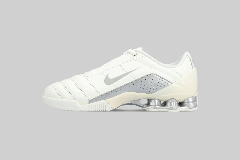 The NIKE T90 SHOX MAGIA QS by Machina - Wearable Technology is a white low-top sneaker with a silver swoosh, silver Shox heel columns, and cuero premium, inspired by the iconic Total 90 line, shown on a plain gray background.