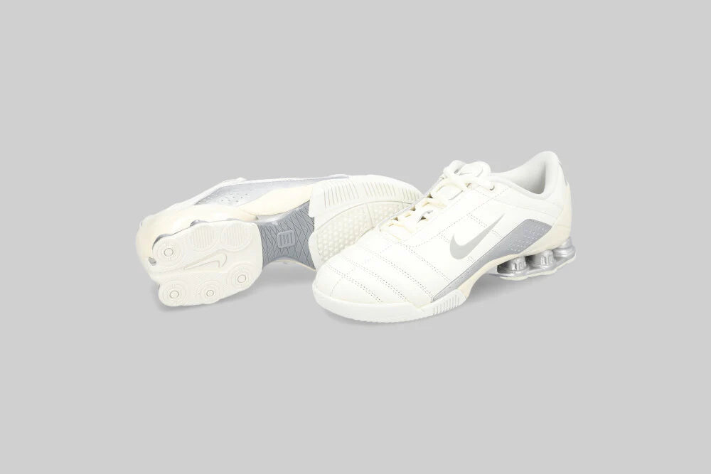 A pair of NIKE T90 SHOX MAGIA 'Sail Metallic Silver' sneakers by Machina - Wearable Technology, with cream leather and silver details, are displayed on a light gray background—one upright, the other showing its sole and Shox technology.