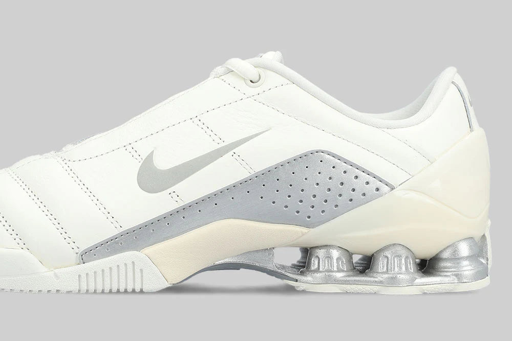 A side view of the NIKE T90 SHOX MAGIA 'Sail Metallic Silver' by Machina - Wearable Technology shows a white sneaker with cream leather, silver swoosh, metallic details, and visible Shox springs in the heel on a gray background.