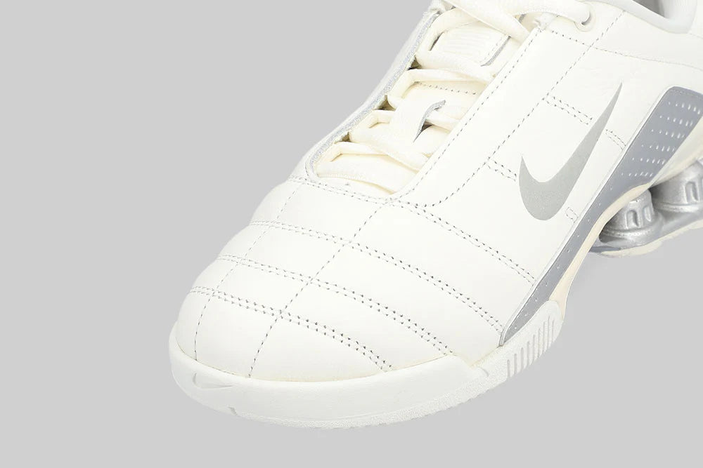Close-up of the NIKE T90 SHOX MAGIA 'Sail Metallic Silver' by Machina - Wearable Technology, a white athletic shoe with silver accents and stitched cream leather details, displayed against a plain gray background.