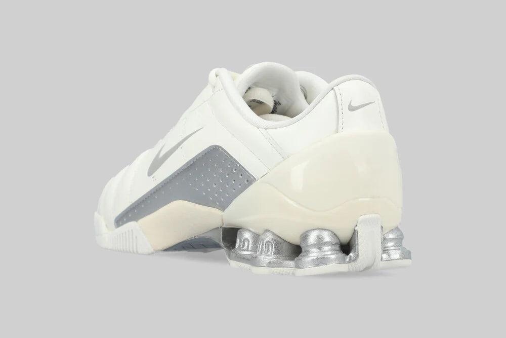 The Machina NIKE T90 SHOX MAGIA 'Sail Metallic Silver' sneaker is displayed from the back and side, highlighting its metallic Shox spring sole and grey swoosh, set against a plain light grey background.