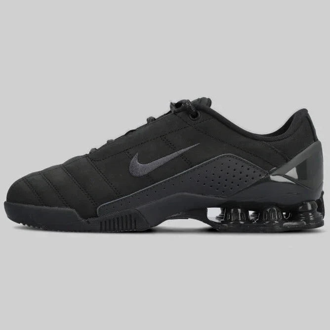 The NIKE TOTAL 90 SHOX MAGIA ‘BLACK’ by Machina - Wearable Technology is a black sneaker with genuine leather, a dark gray Swoosh logo, visible Shox tech in the heel, and a low-cut design on a light gray background.