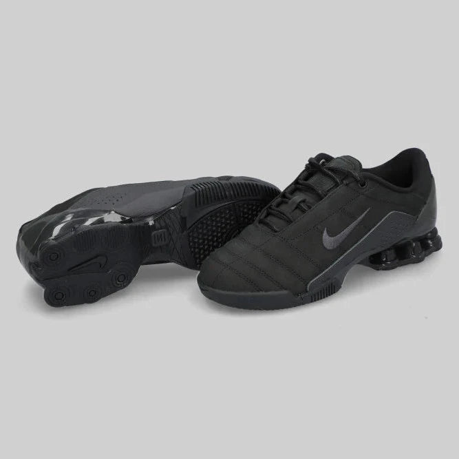 A pair of NIKE TOTAL 90 SHOX MAGIA ‘BLACK’ by Machina - Wearable Technology, in black with a gray swoosh, genuine leather, shown with one shoe upright and the other on its side to reveal sole and heel details.