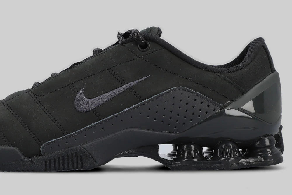 The NIKE TOTAL 90 SHOX MAGIA ‘BLACK’ by Machina - Wearable Technology is a sleek black low-top sneaker with signature Shox heel columns and the Nike swoosh, shown on a plain gray background.