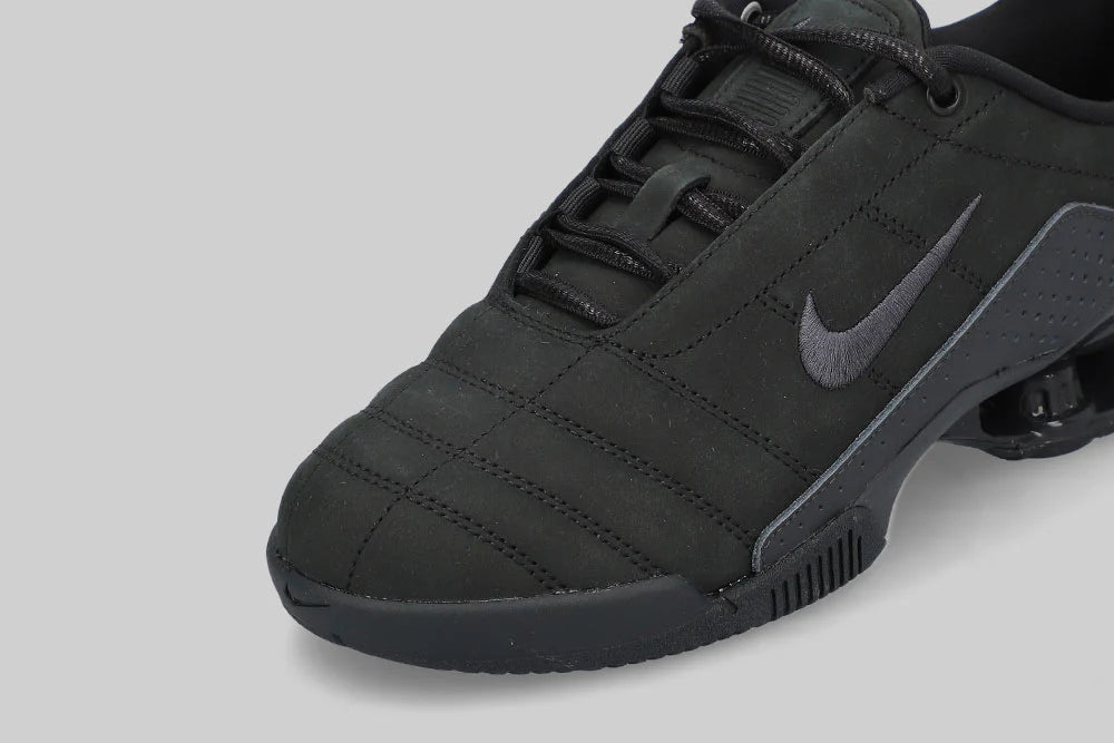 Close-up of the NIKE TOTAL 90 SHOX MAGIA ‘BLACK’ by Machina - Wearable Technology, showing genuine black leather, prominent Swoosh logo, textured stitching, and a partly visible air-cushion sole on a light gray background.