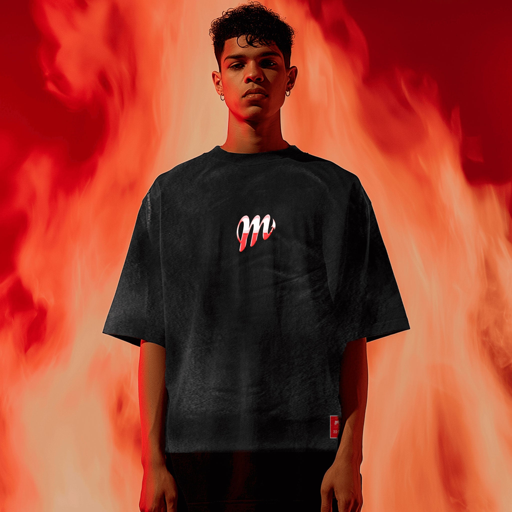 Against a blazing red and orange backdrop, an individual dons the (Copy) Diablos Isotype Chromed black T-shirt from Machina - Wearable Technology, featuring a sleek M logo. Their relaxed demeanor stands in contrast to the fiery background, capturing the spirit of loyalty and fervor for Diablos Rojos del México.