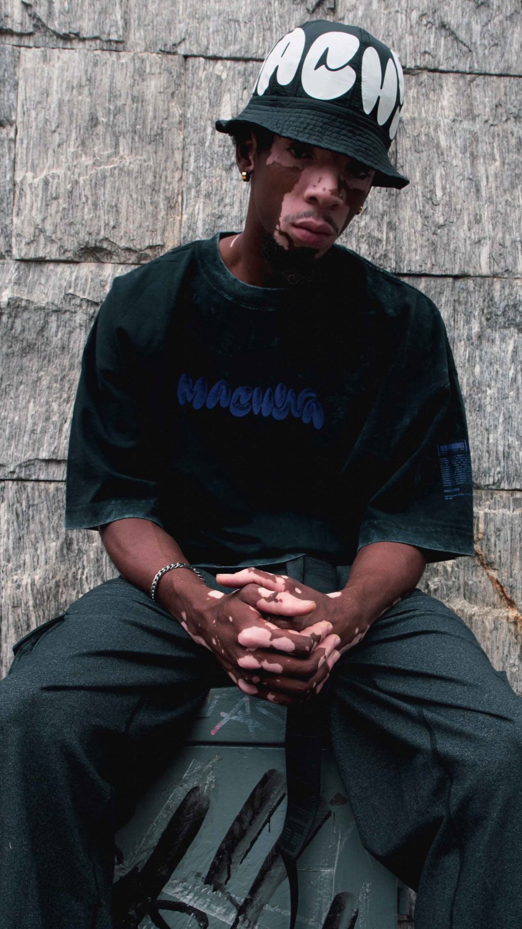 A person wearing a loose black shirt from Machina's Spine collection and a bucket hat adorned with bold white lettering sits against a textured stone wall. With their hands clasped, they gaze down in contemplation, seemingly pondering the mysteries of cryptid science.