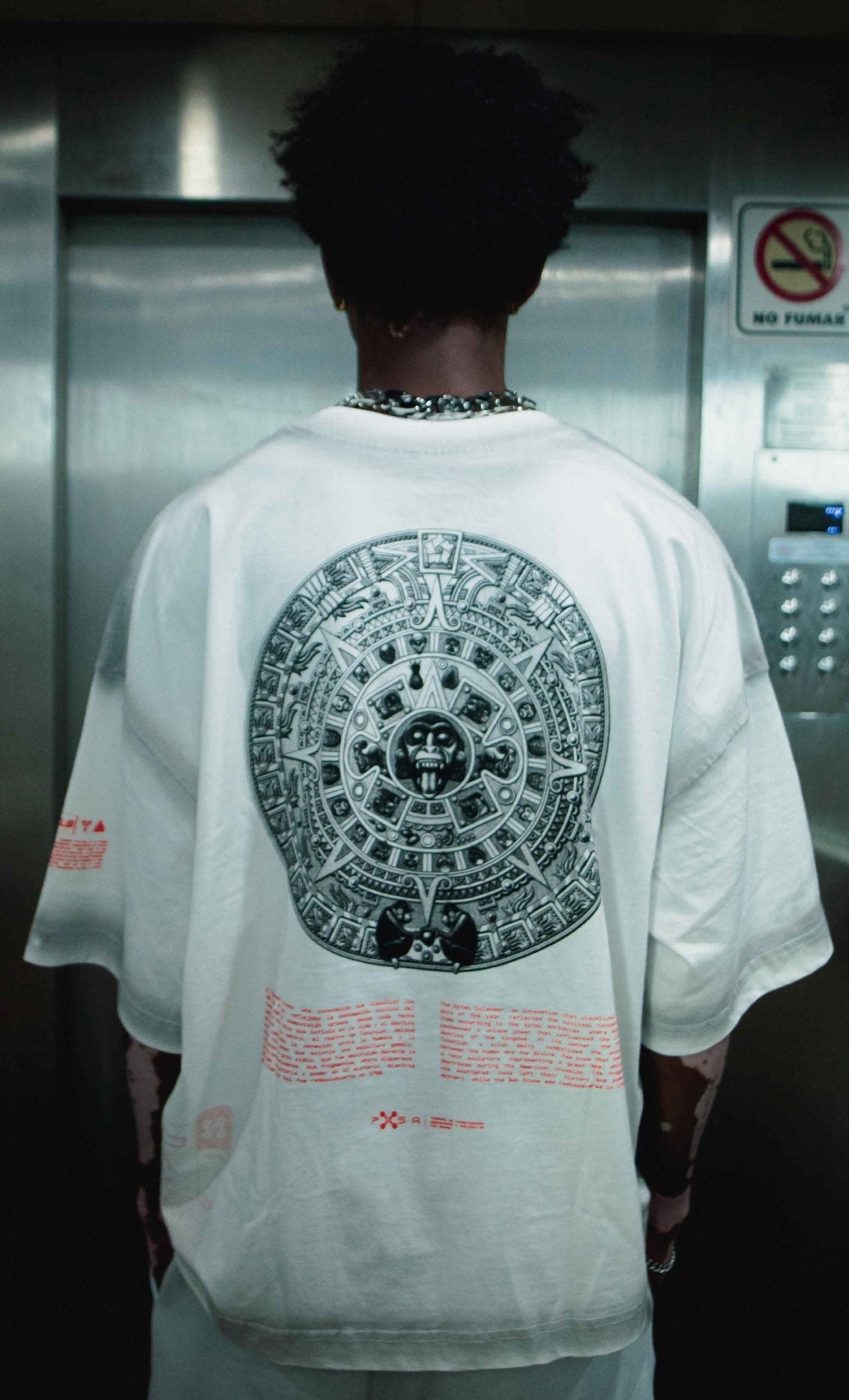 In a metallic-walled elevator, a person dons an oversized white T-shirt from Machina - Wearable Technology, featuring the intricate circular design of the **Calendario Pie Azteca** on the back, evoking an air of ancient power.