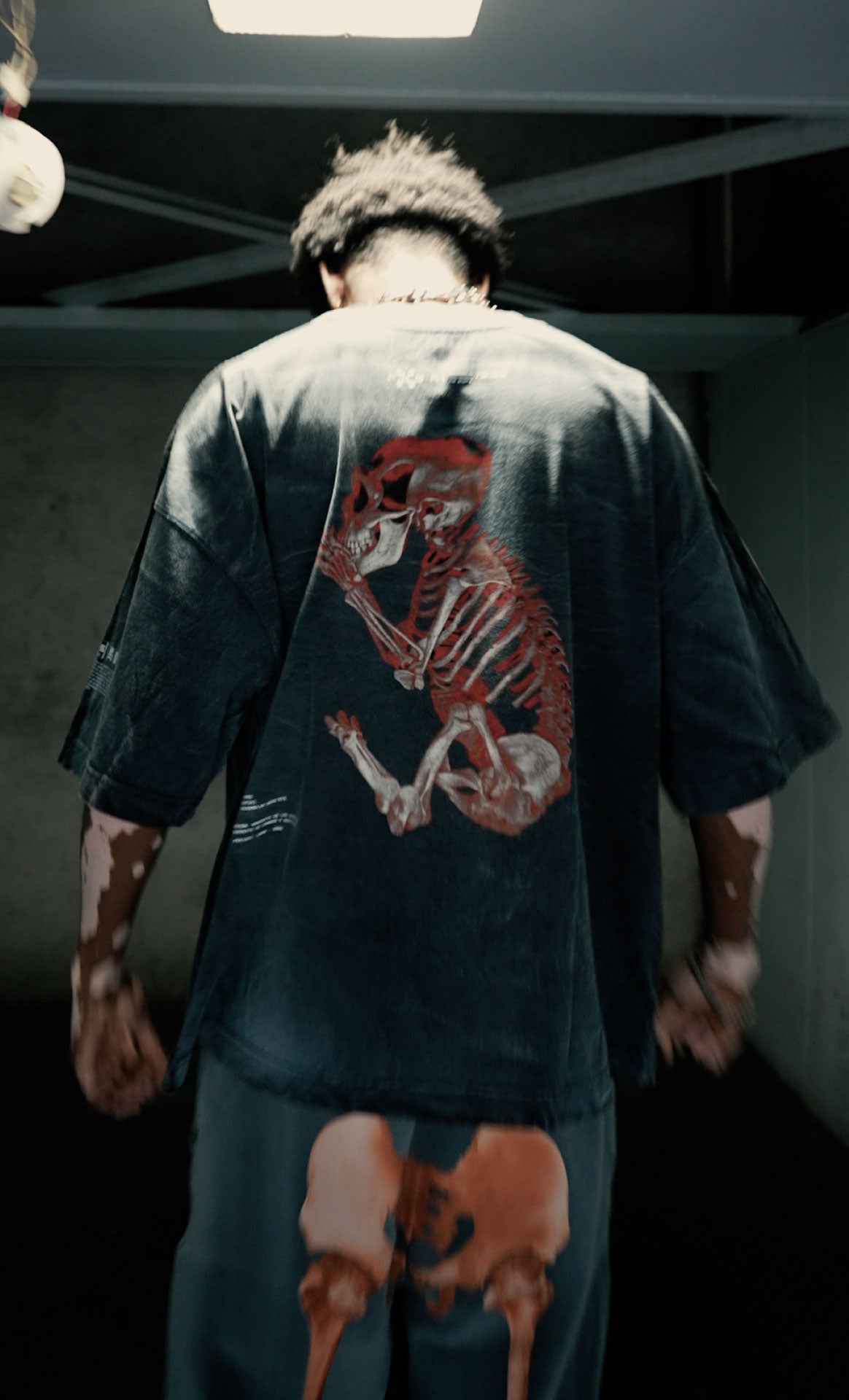 A person stands with their back to the camera, wearing a dark shirt adorned with The Bigfoot Project red skeletal monkey print by Machina - Wearable Technology. In dim light, a smaller mirrored design appears on their pants, hinting at biomechanical hybridization.